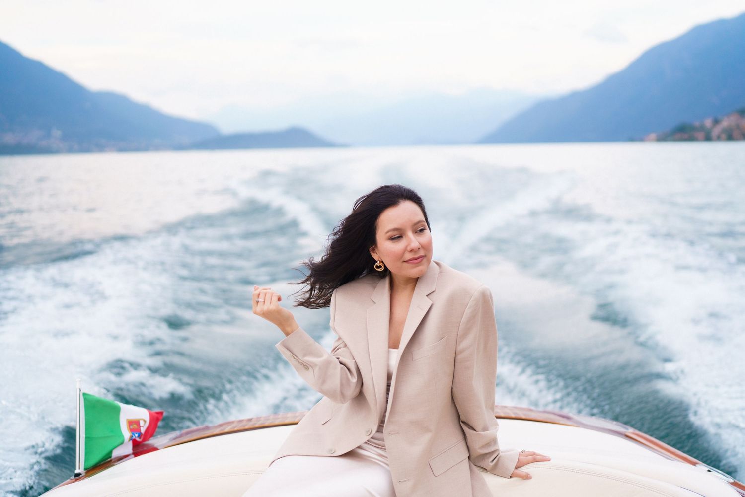 Person enjoying a boat ride on a lake with mountains in the background, wearing a beige blazer.