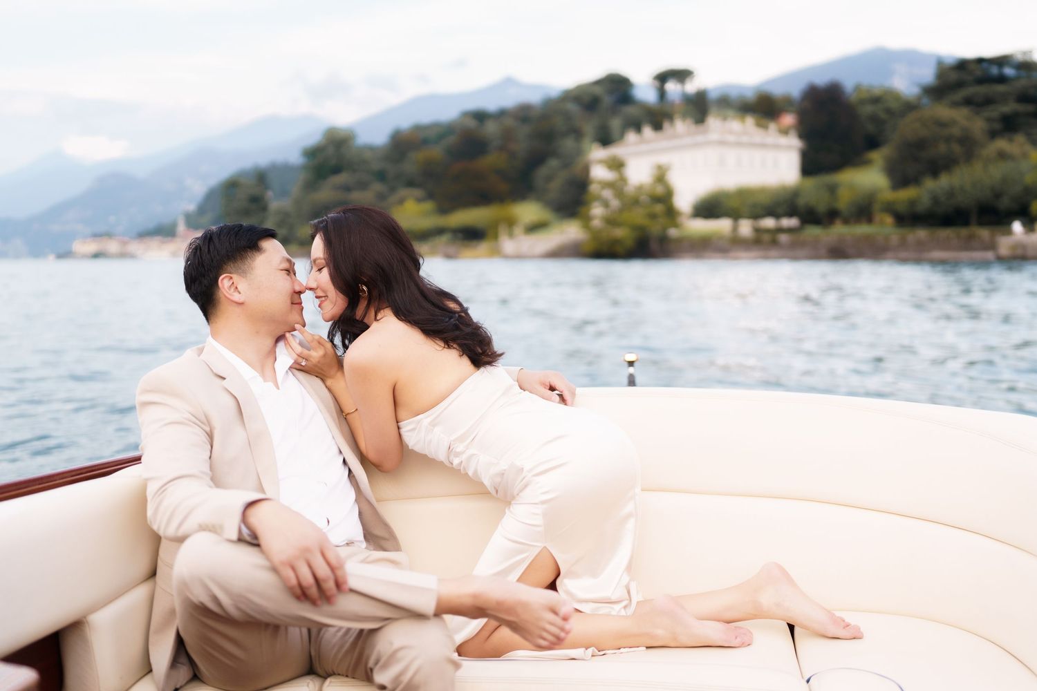 A romantic couple in light colored outfits sits together on a boat at sunset by the ocean.