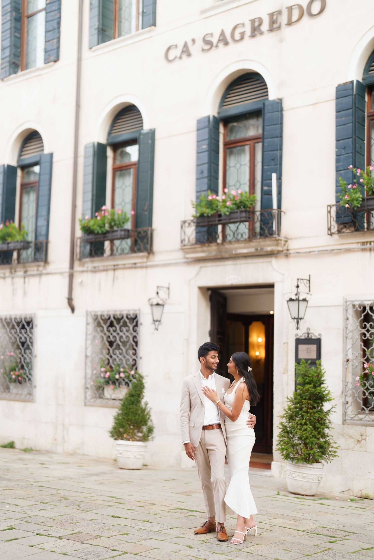 A couple stands outside the elegant Ca'Sagredo hotel entrance in Venice with ornate architecture and flower boxes.