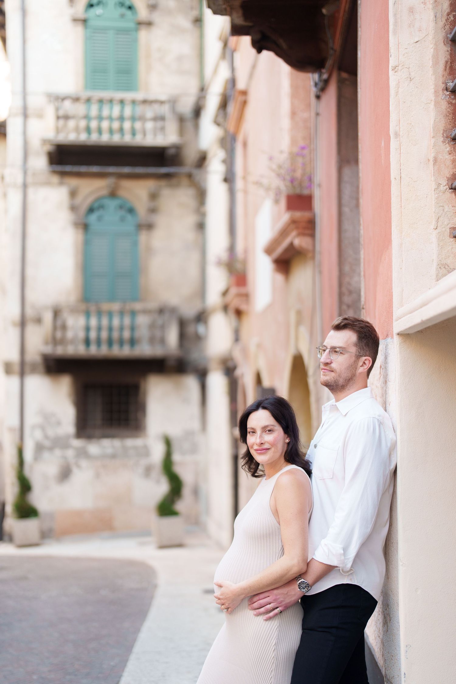 Couple in white and black attire embracing in romantic poses against pink Mediterranean building walls.