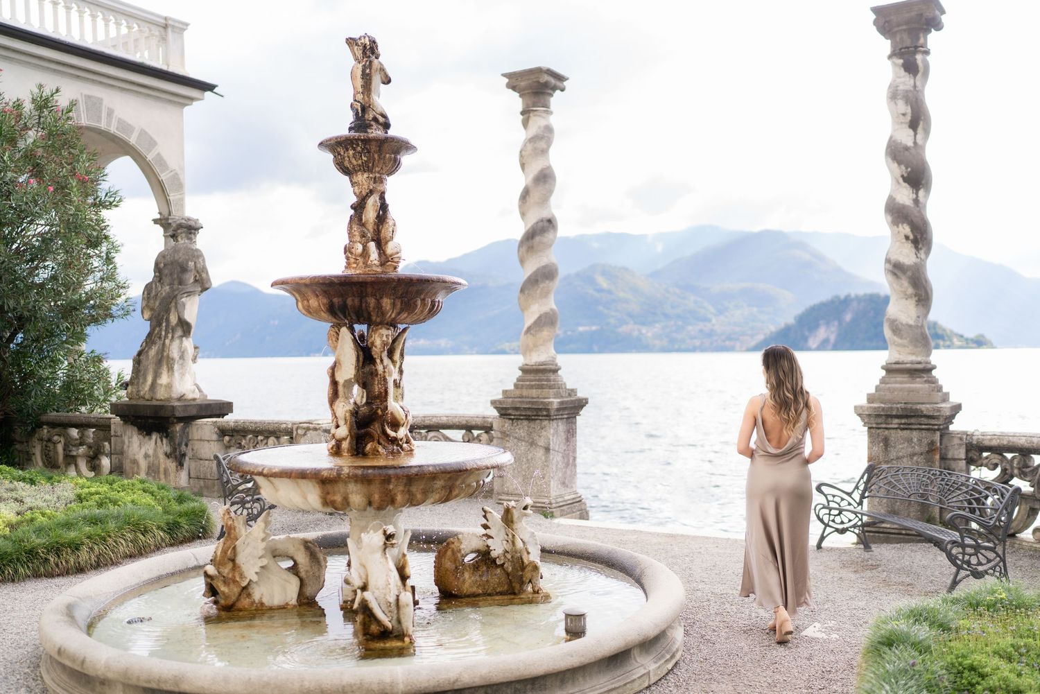 A scenic fountain with ornate sculptures stands near a lake with mountains in the background at Lake Como, Italy.