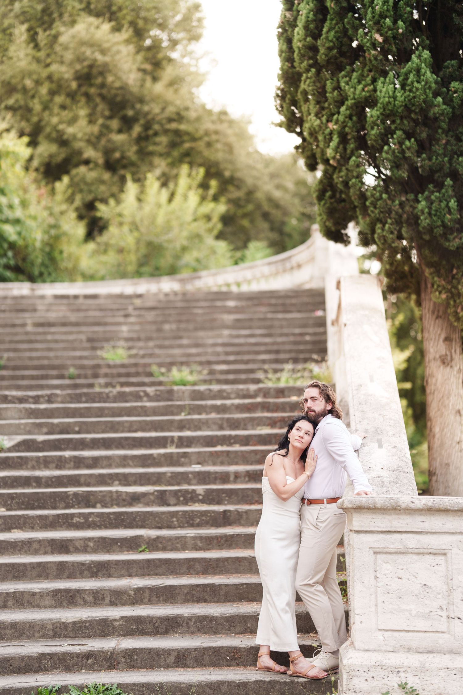 A romantic couple embraces on stone steps in an outdoor garden setting with trees in the background.