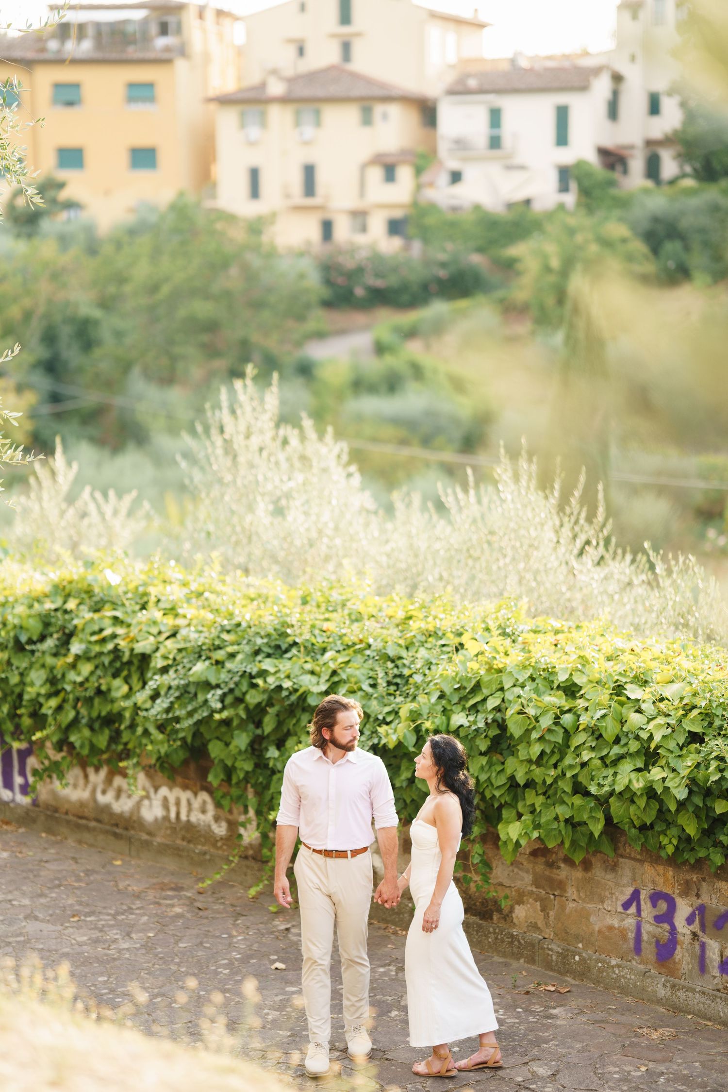 A romantic couple strolls along a stone wall with lush greenery and Italian villas in the background.