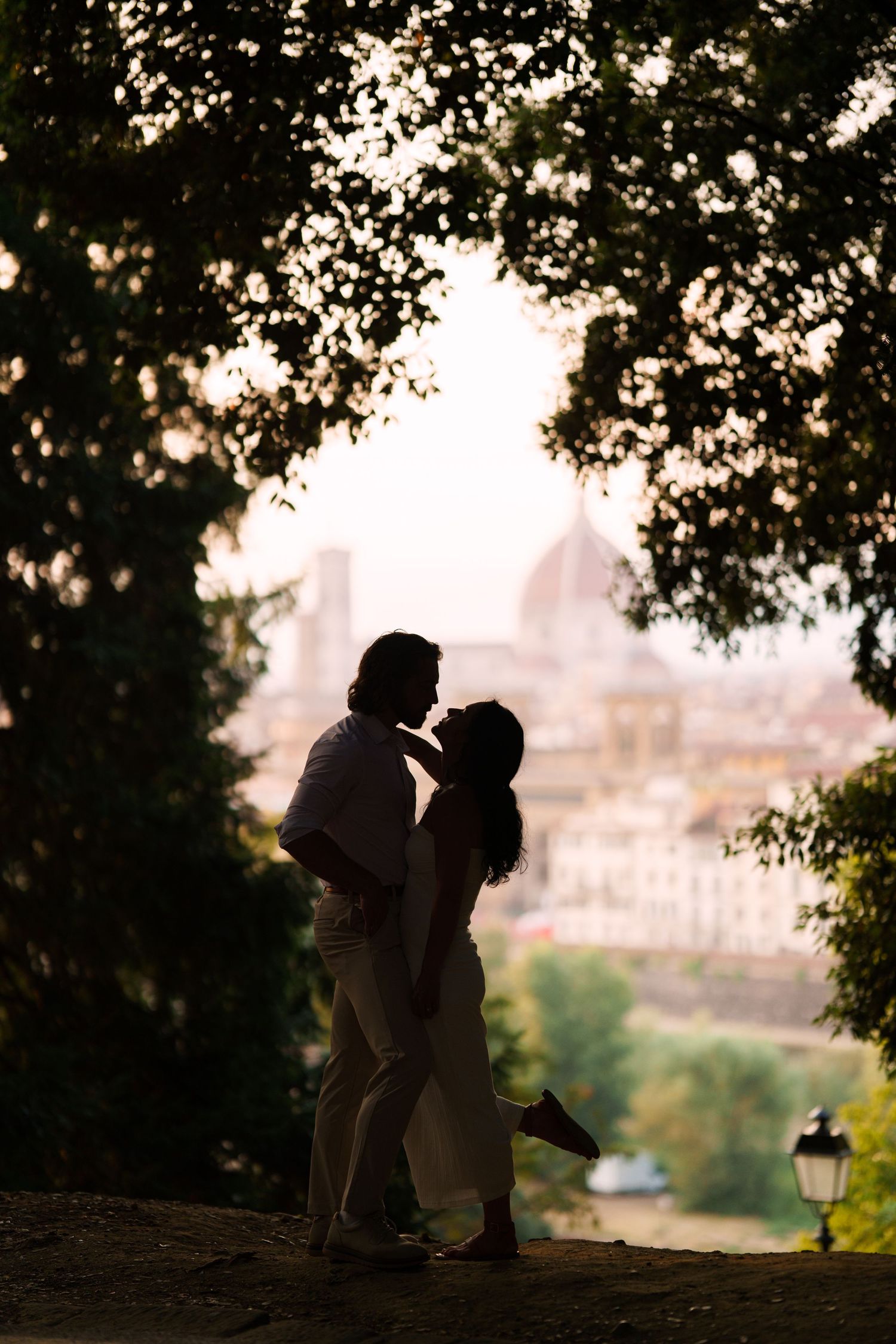 Silhouetted couple sits together on a hill overlooking a city at sunset through tree branches.