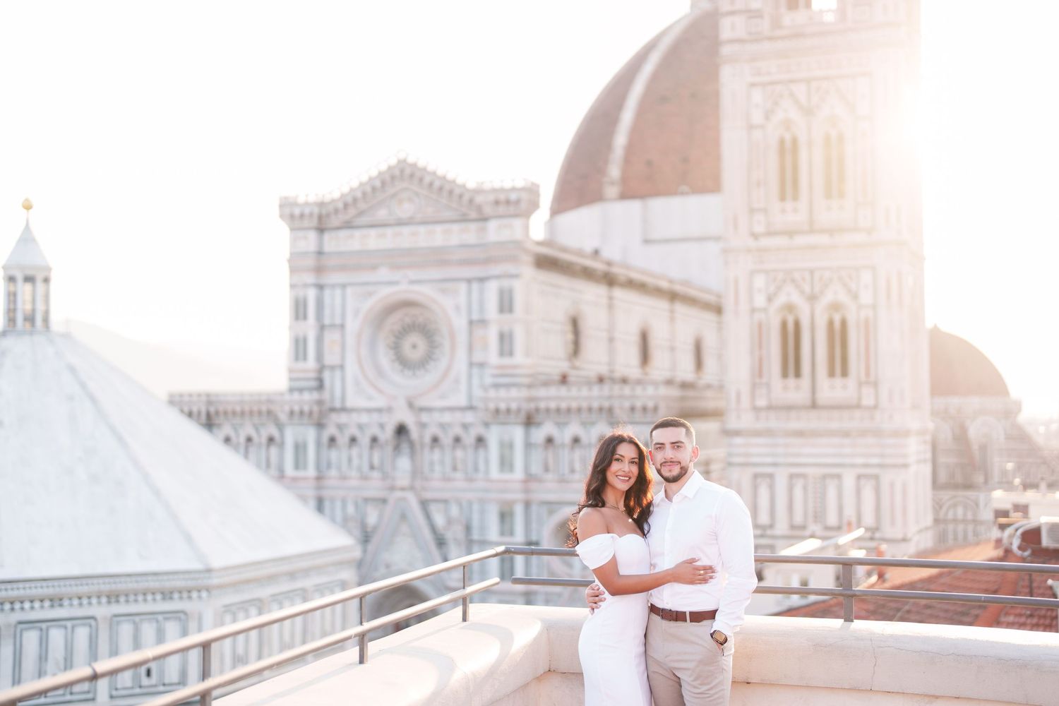 Couple in white outfits embracing on rooftop terrace with Florence Cathedral and Duomo in background during sunset.