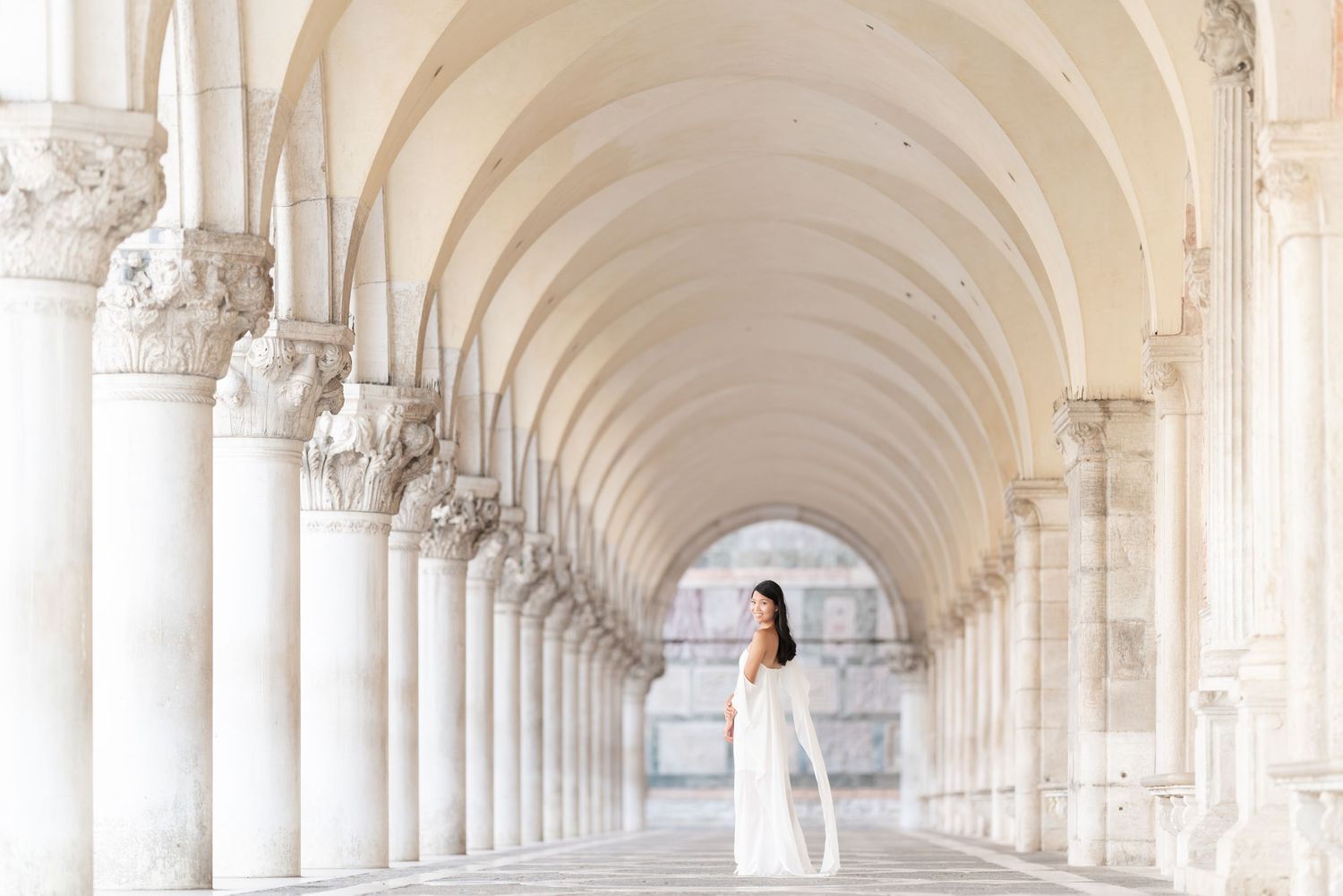 A person in a white dress stands in an elegant arched colonnade with ornate columns stretching into the distance.