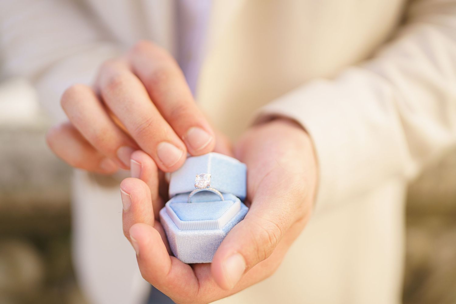 A close-up series of hands holding a small engagement ring box in various poses against a blurred background.