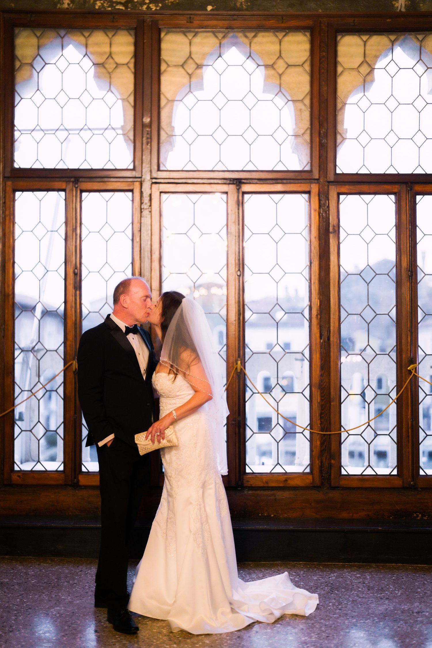 A romantic wedding couple dances in a grand room with ornate leaded glass windows creating a timeless silhouette.