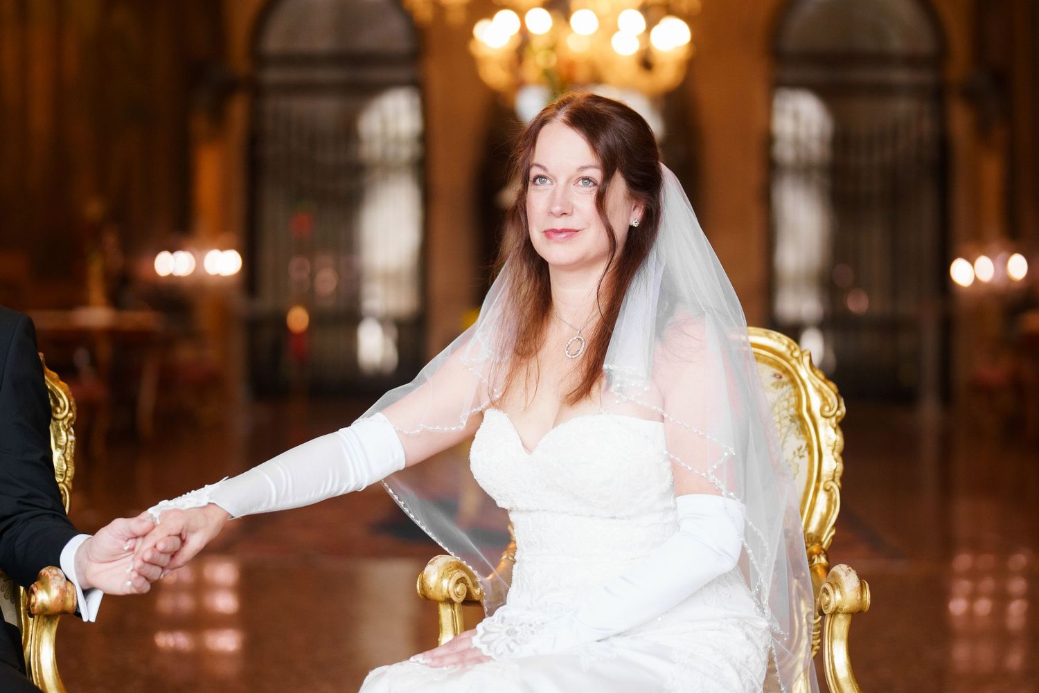 A bride in a white wedding gown and veil poses in an elegant venue with ornate chandeliers and golden decor.