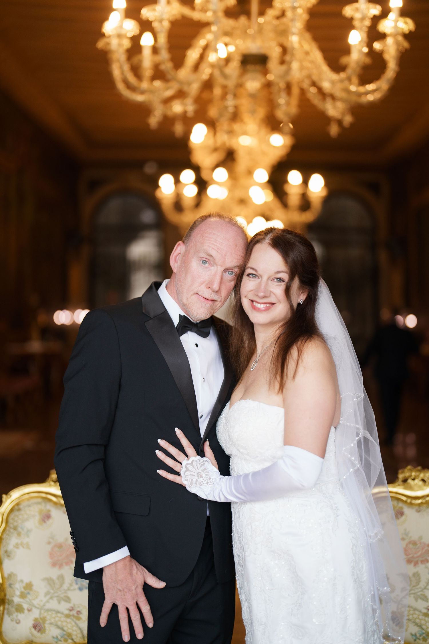 Romantic wedding couple poses together in an elegant ballroom with ornate crystal chandeliers glowing warmly above.