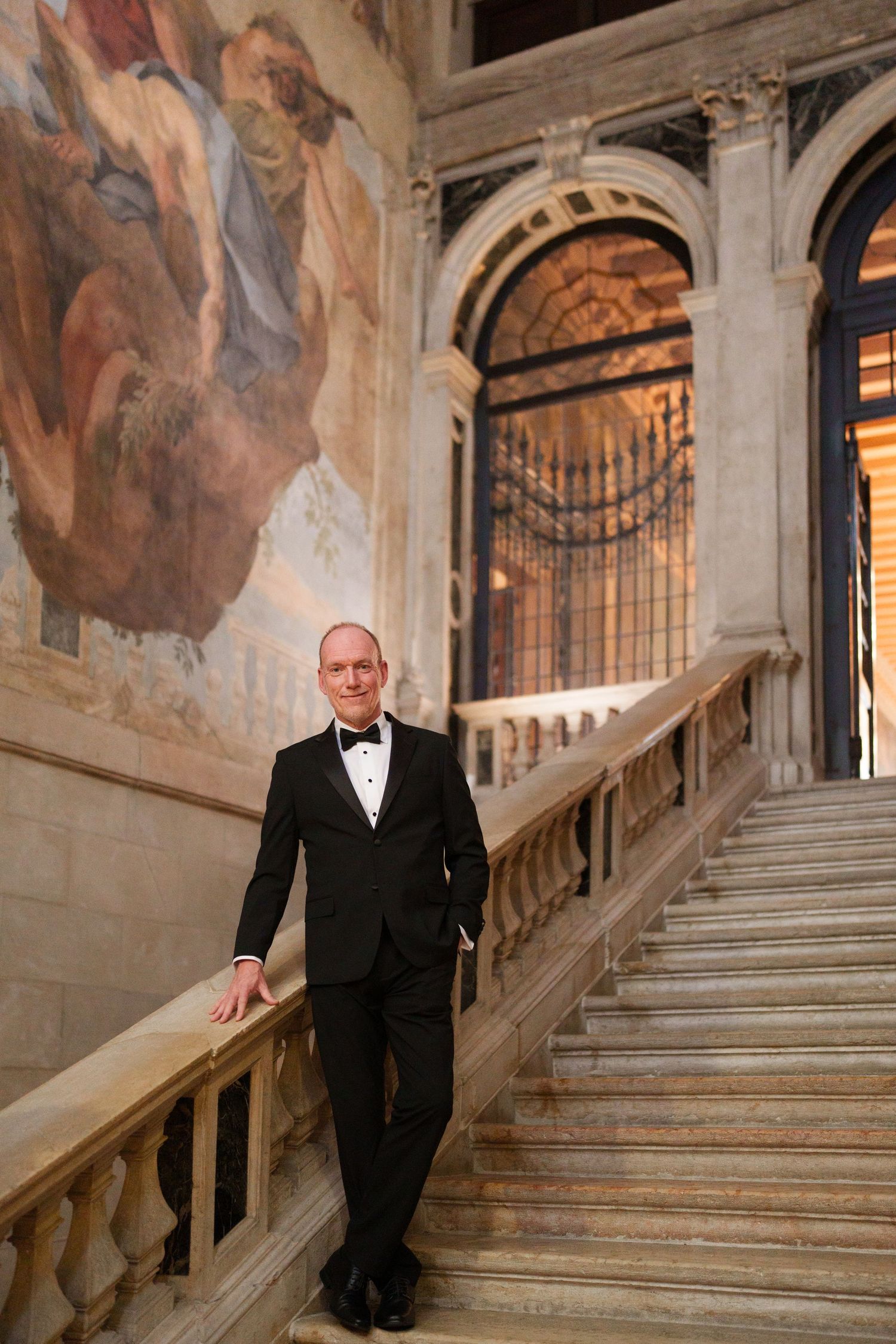 A person in a black suit poses on an ornate marble staircase with decorative frescoes and classical architecture.