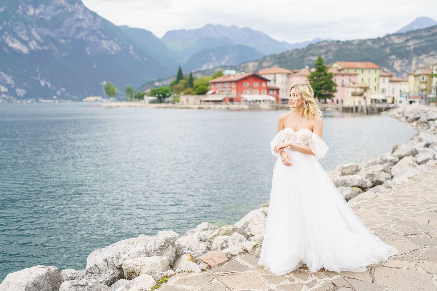Wedding couple poses on rocky shoreline of Lake Garda with Italian mountains and historic buildings in background.