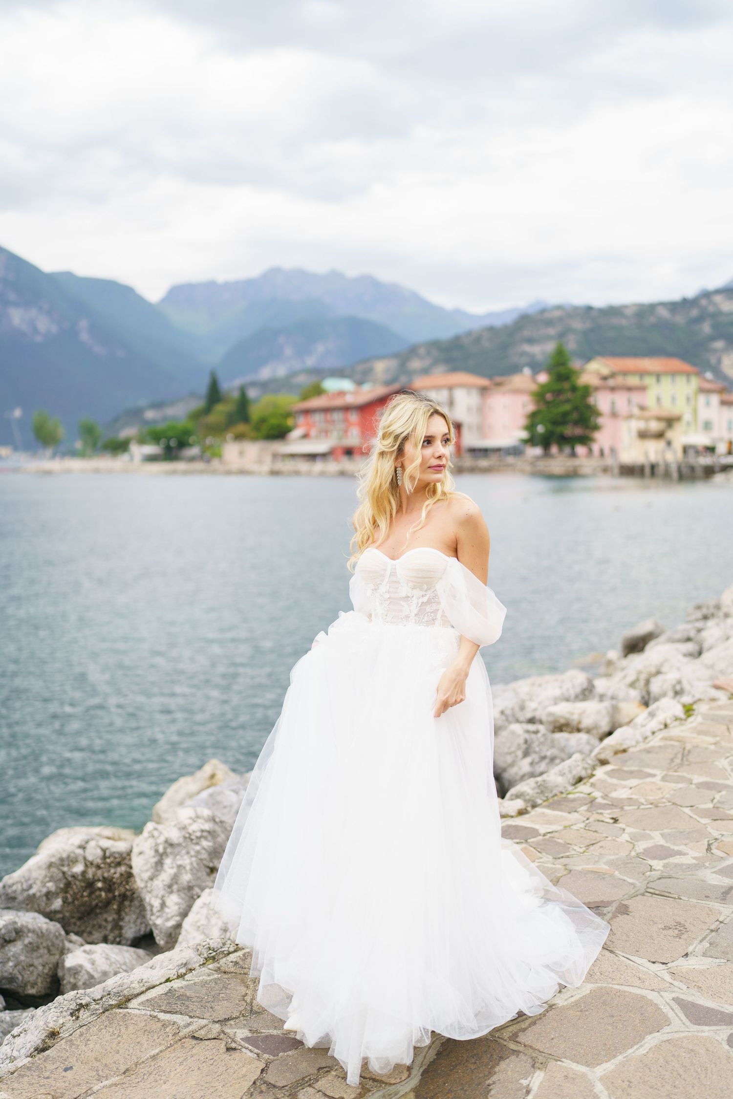A white wedding dress flows elegantly along rocky coastal shores with mountains and buildings in the background.