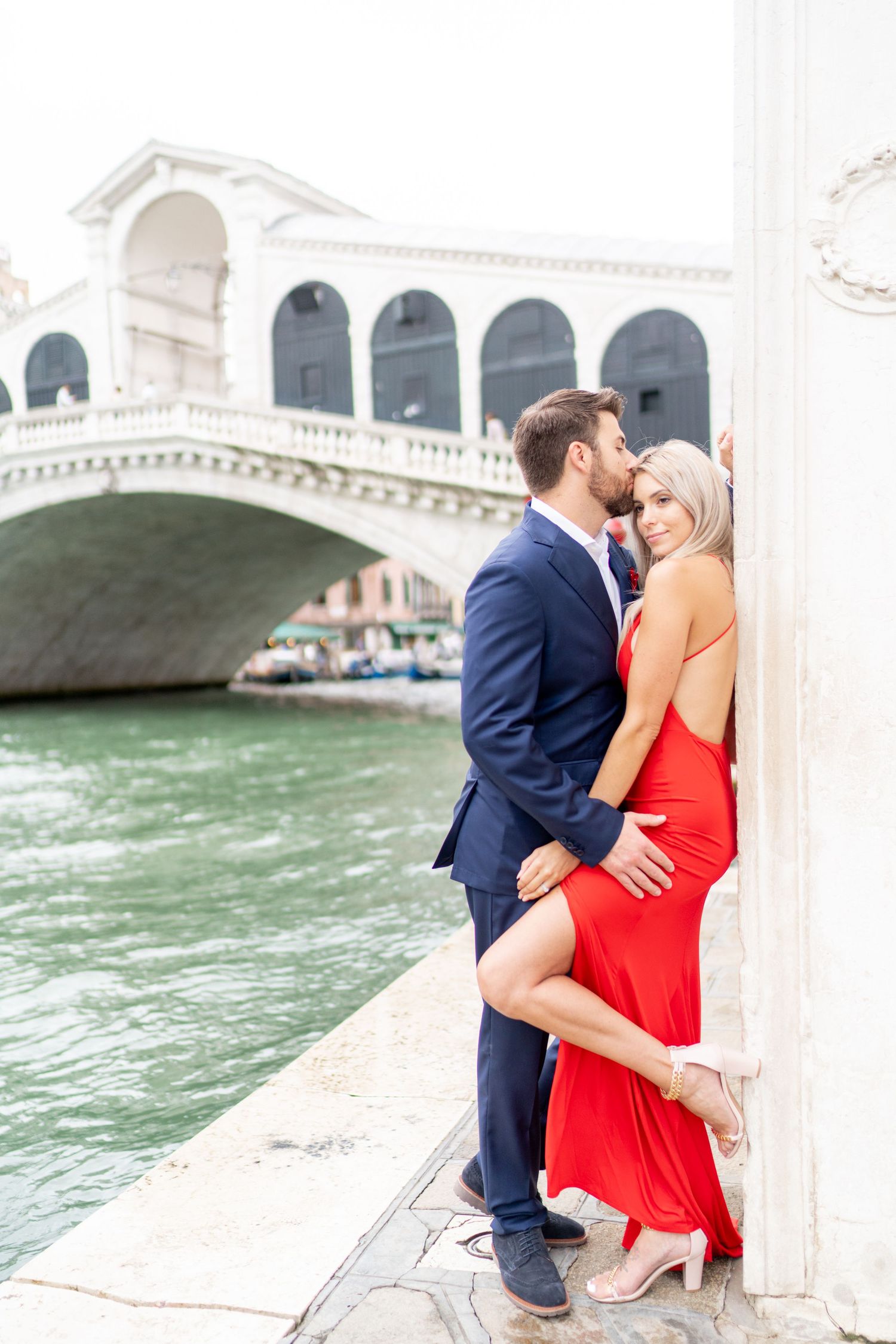 Romantic couple embracing by the Rialto Bridge in Venice, Italy with turquoise canal waters in the background.