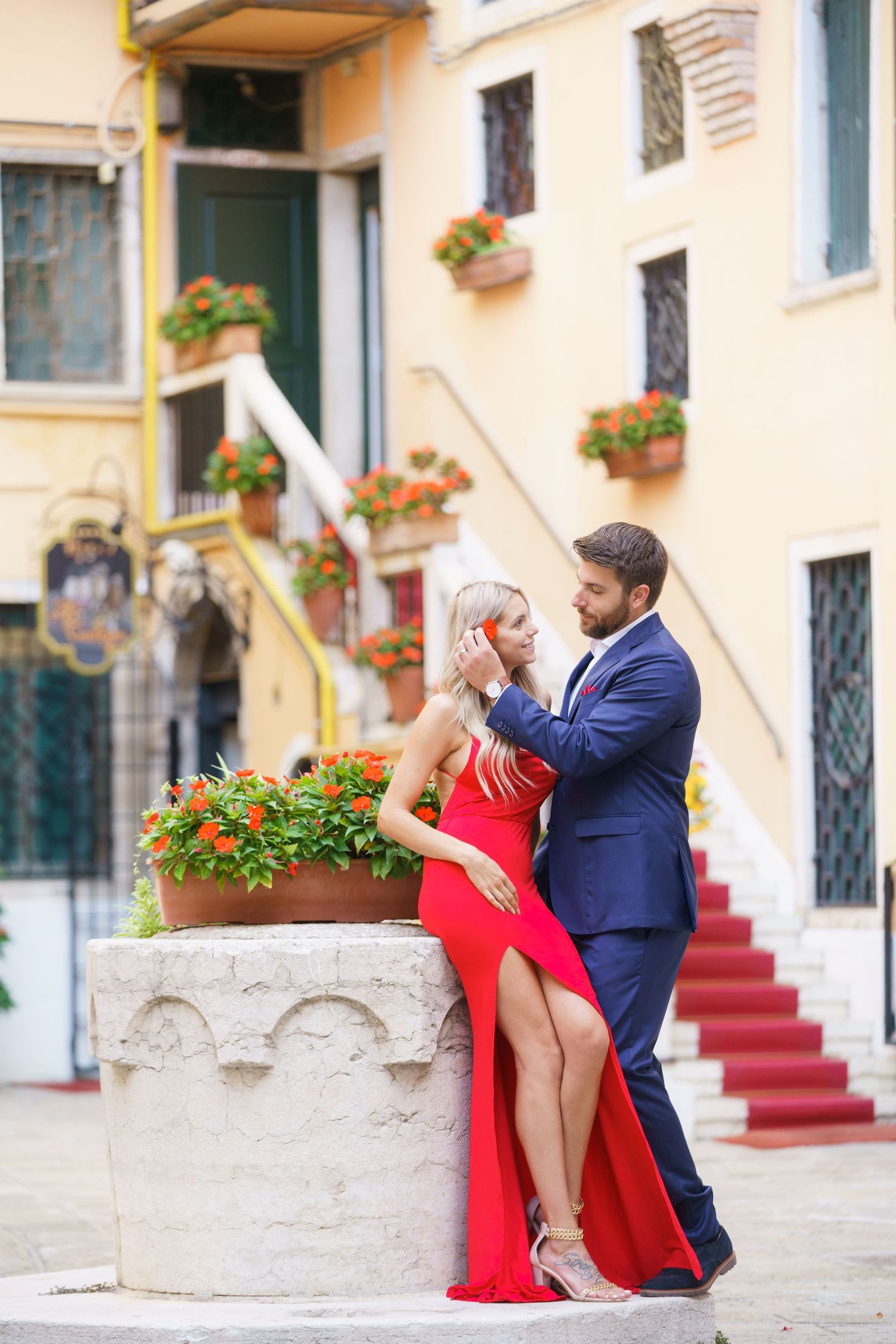 Romantic couple in red dress and navy suit embrace on stone steps with flower planters in European setting.