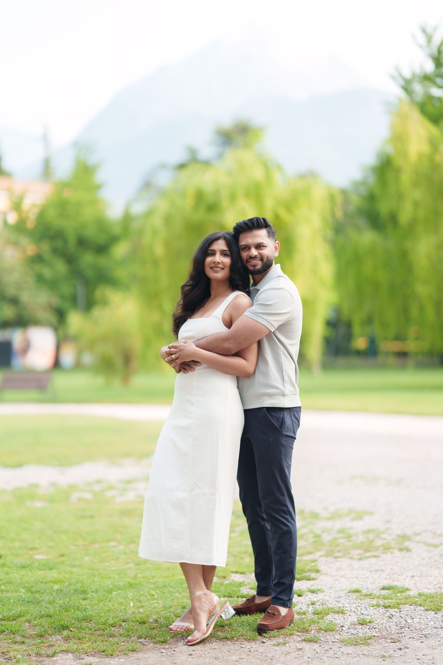 A couple embraces while standing on a gravel path surrounded by green trees and grass in a park setting.