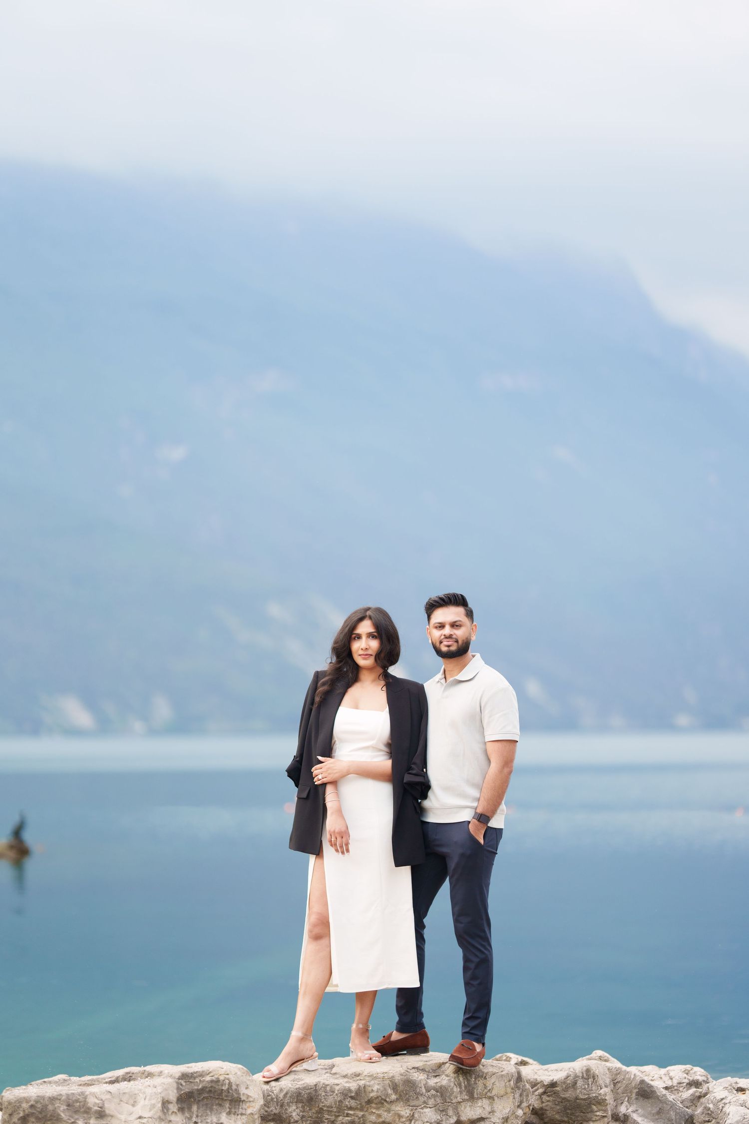 Couple standing on rocky overlook with scenic ocean and mountain views in the background.