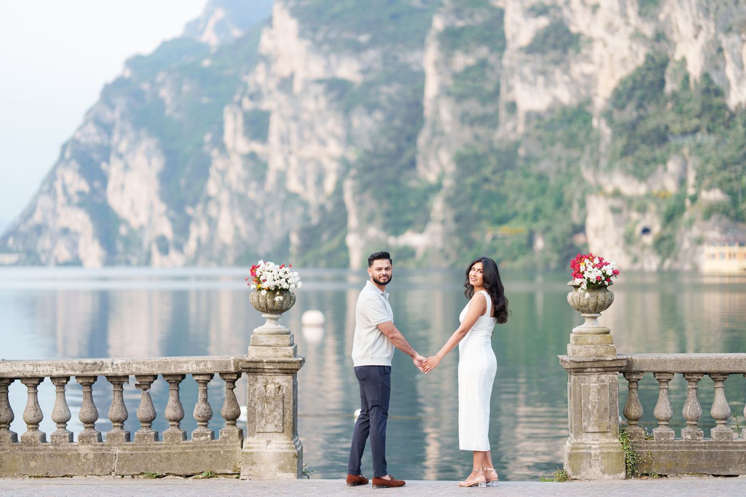 A couple poses romantically on a stone balcony overlooking a scenic lake and mountain landscape.