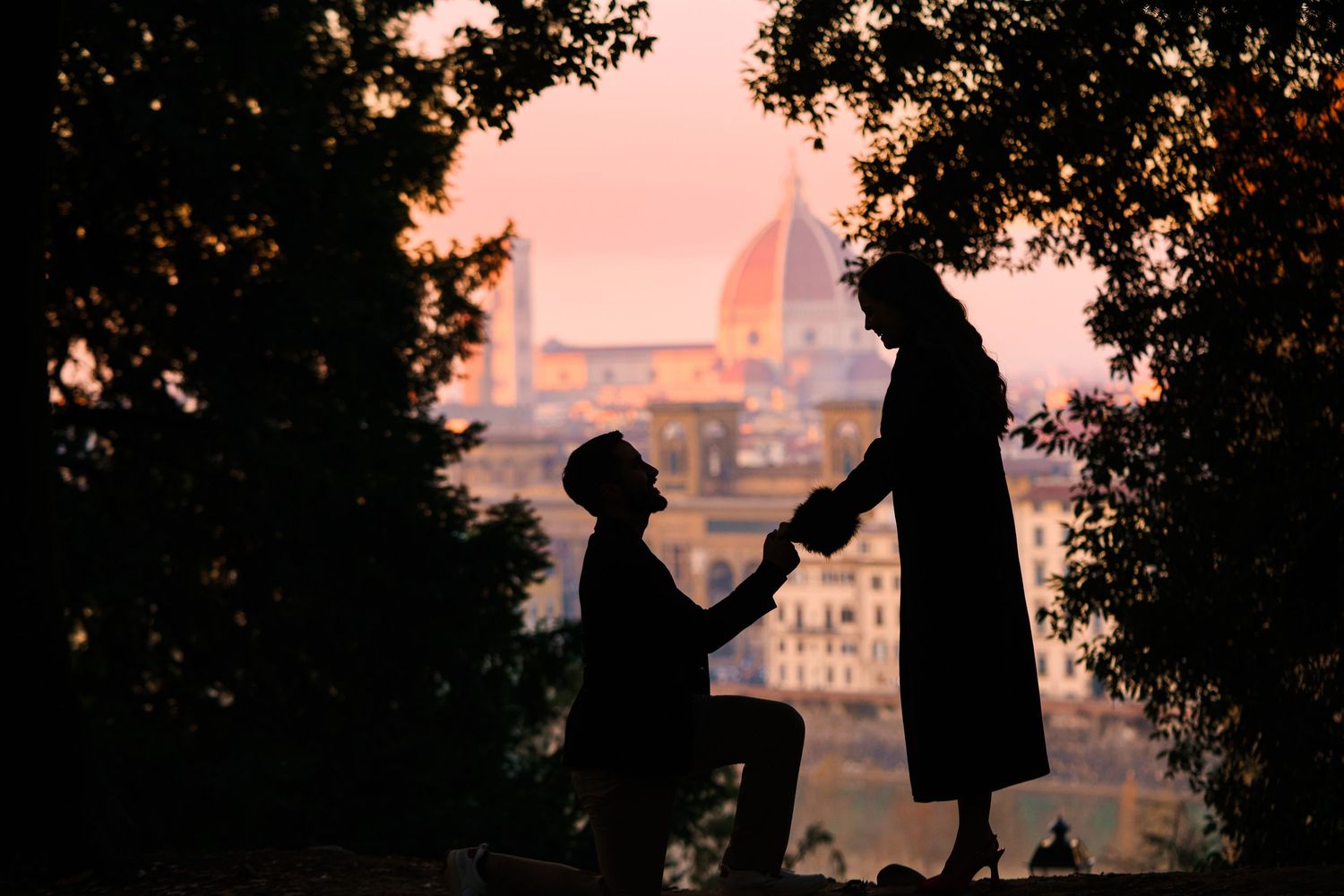 Silhouetted figures walk along a path at sunset with trees and buildings in the background.