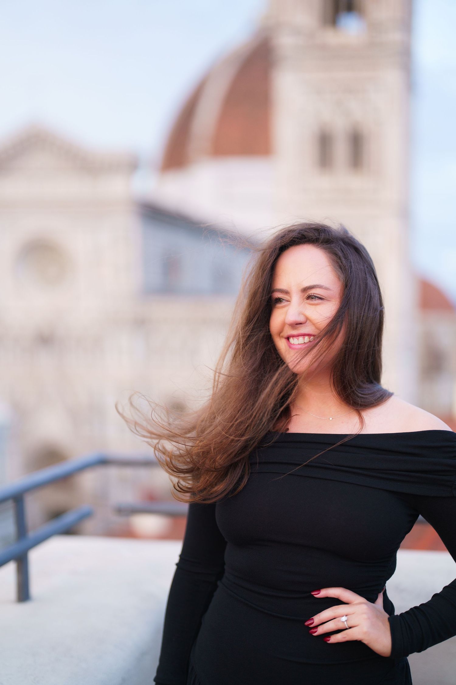 Woman in black off-shoulder dress smiling with windswept hair against backdrop of Florence Cathedral in Italy.
