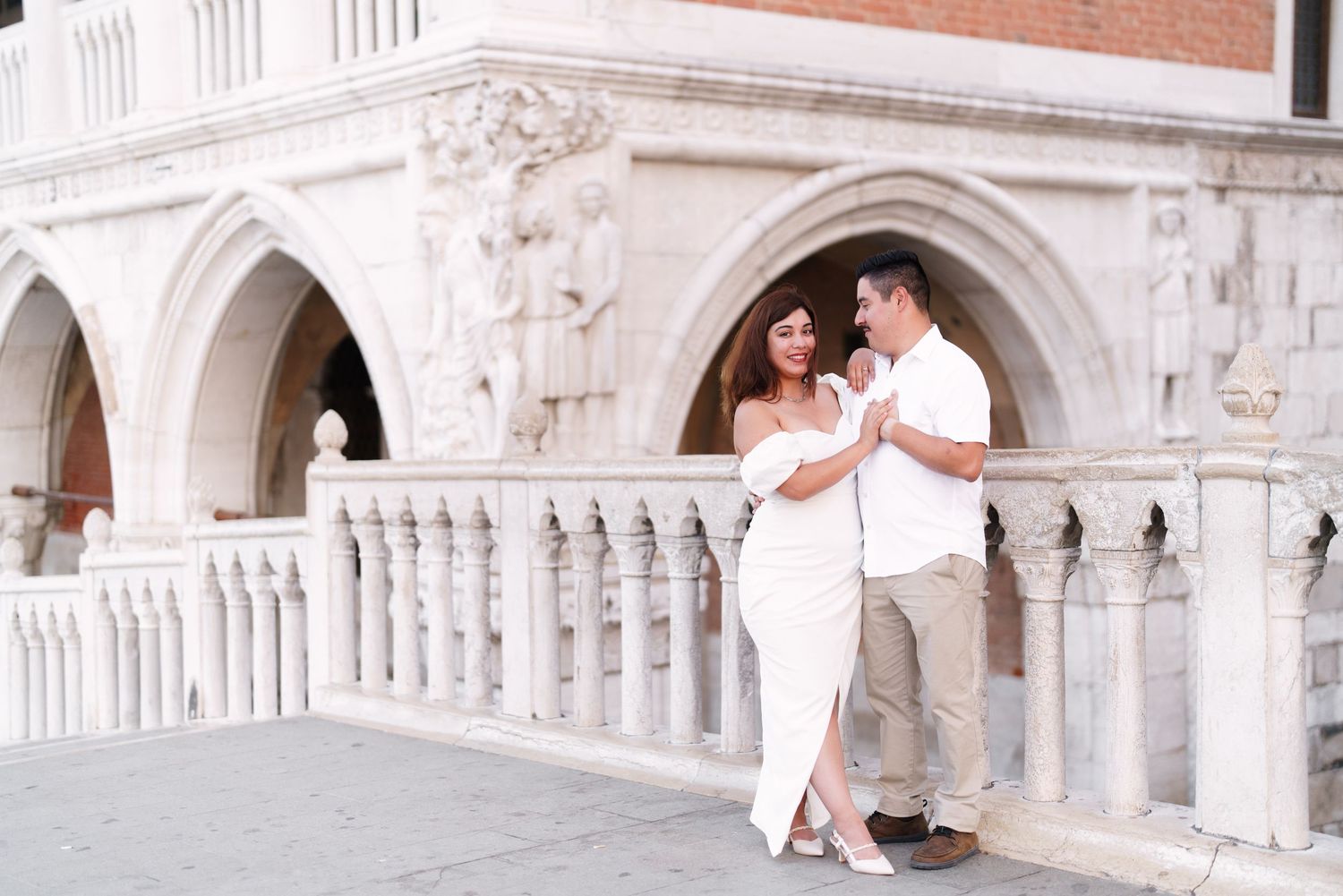 A couple in white outfits shares a romantic moment on an ornate marble balcony with arched architecture in Venice.