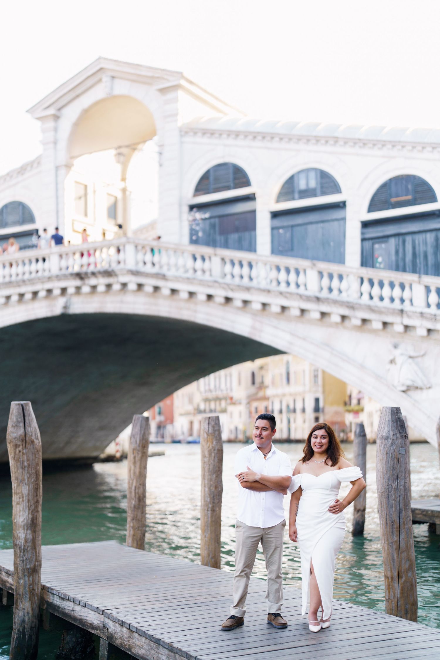 Couple standing on wooden dock near iconic Rialto Bridge in Venice with grand white arches and canal in background.