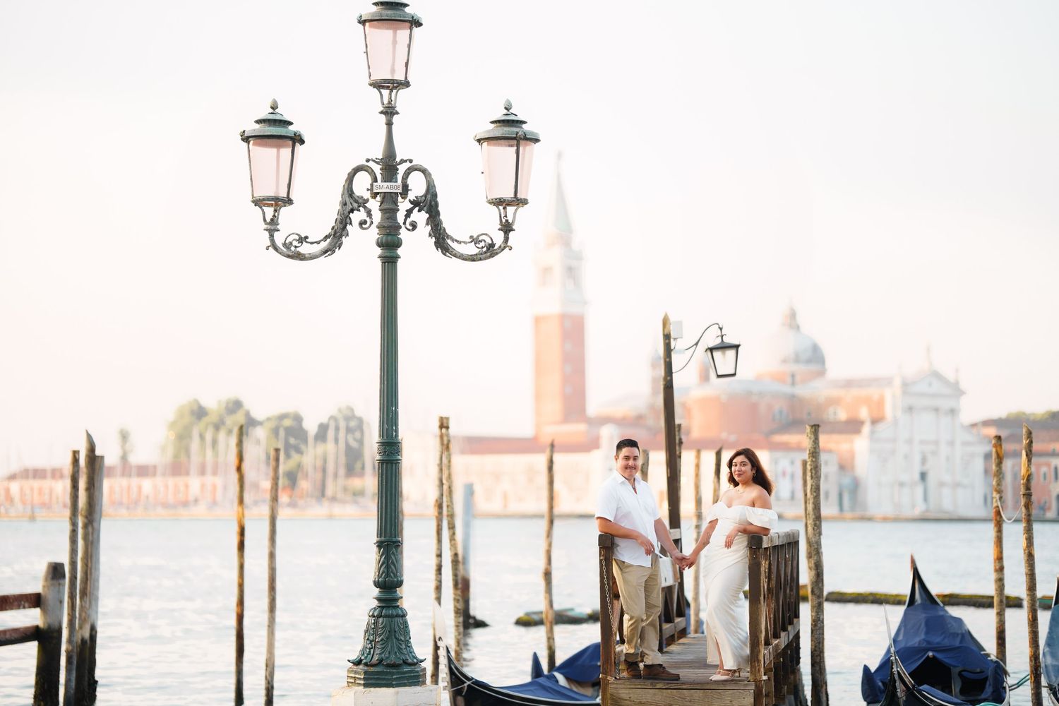 Venetian gondolas moored near ornate lamp posts along a canal with historic buildings and the lagoon in the background.