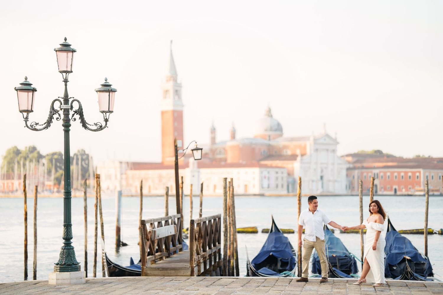 Traditional Venetian lamp posts and covered gondolas line the waterfront with San Giorgio Maggiore church in the distance.