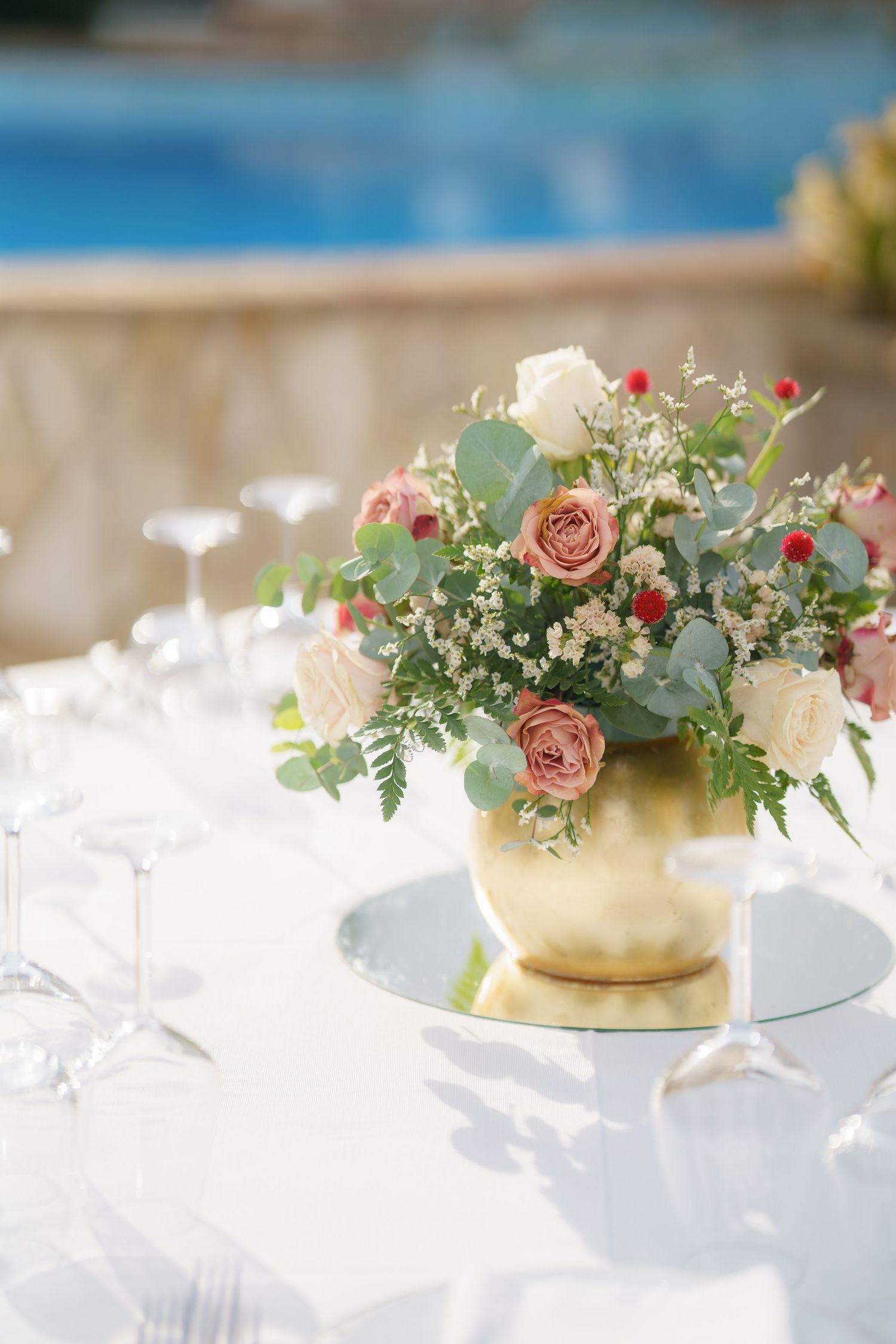 Elegant table centerpieces featuring roses and eucalyptus in gold vessels on white linens.