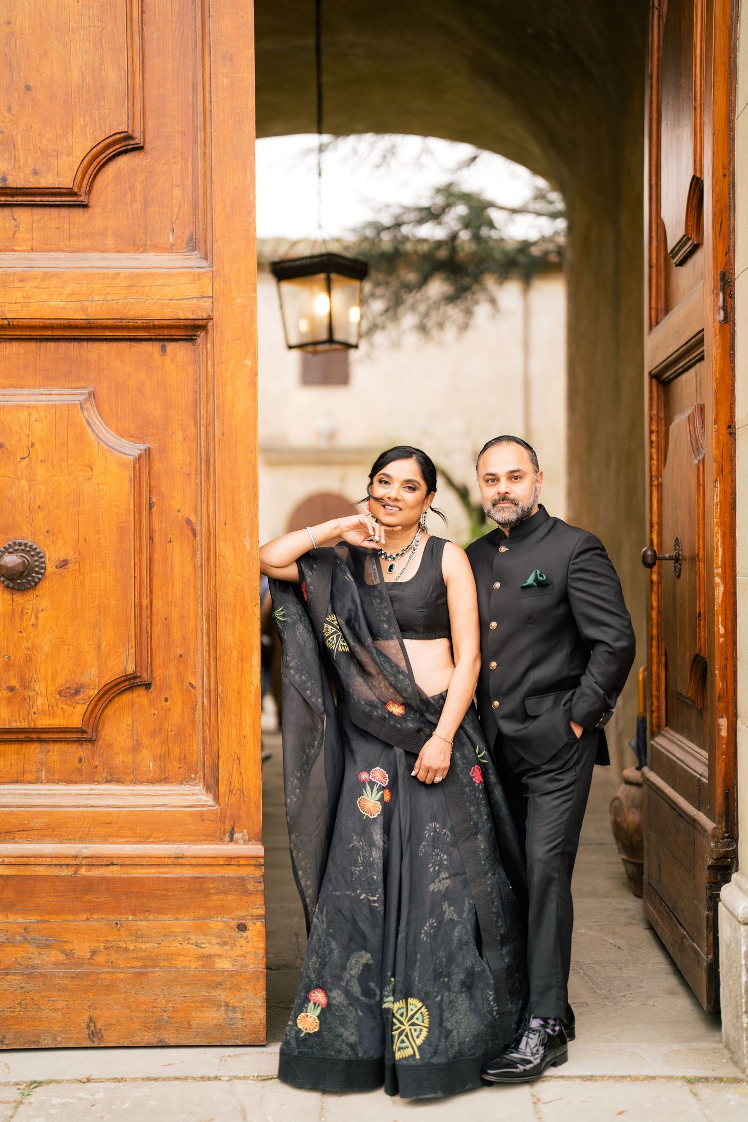 Couple in black attire pose together under arched entryway with vintage wooden door.