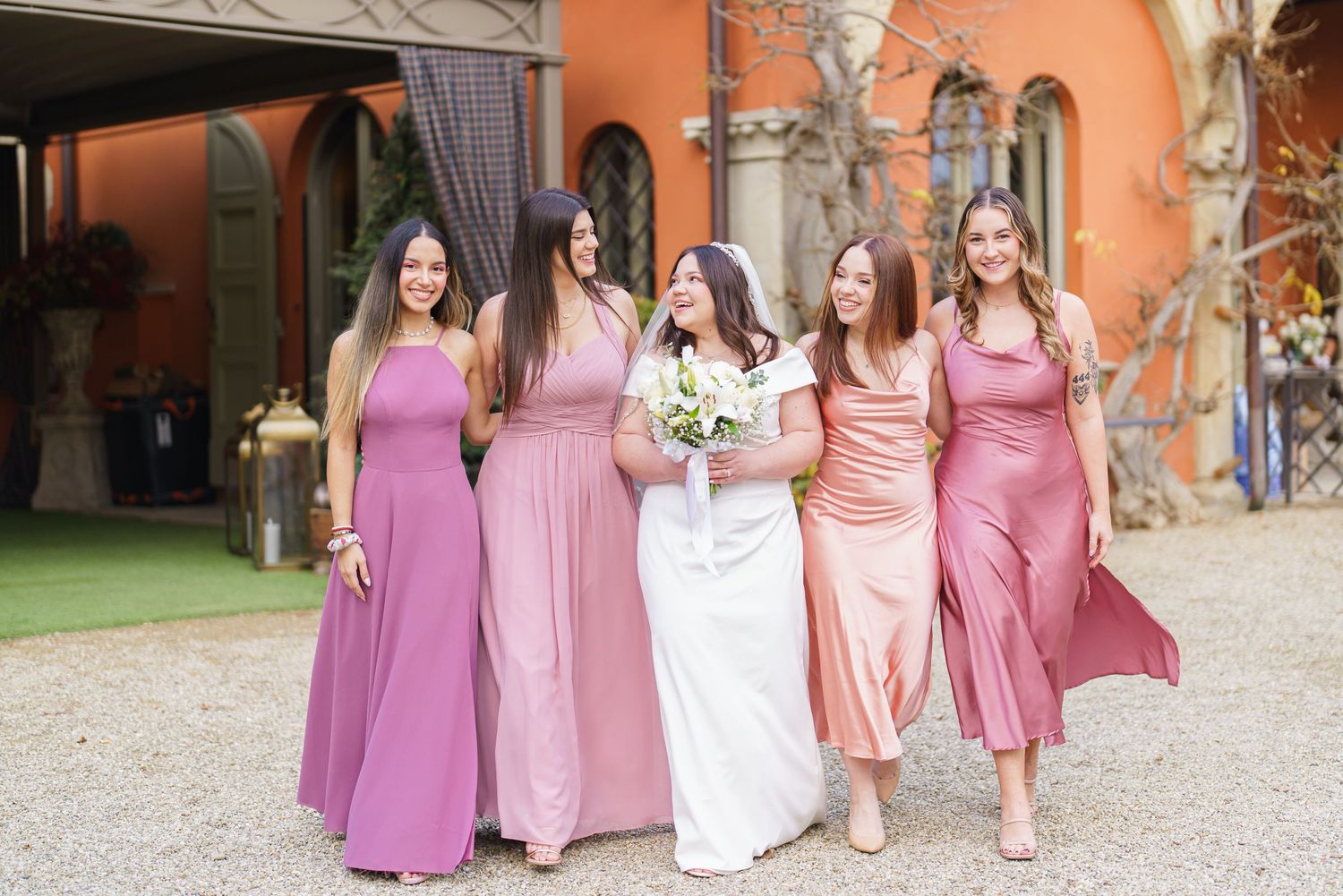 Wedding party in pink and purple dresses poses in front of terra cotta colored Spanish architecture.