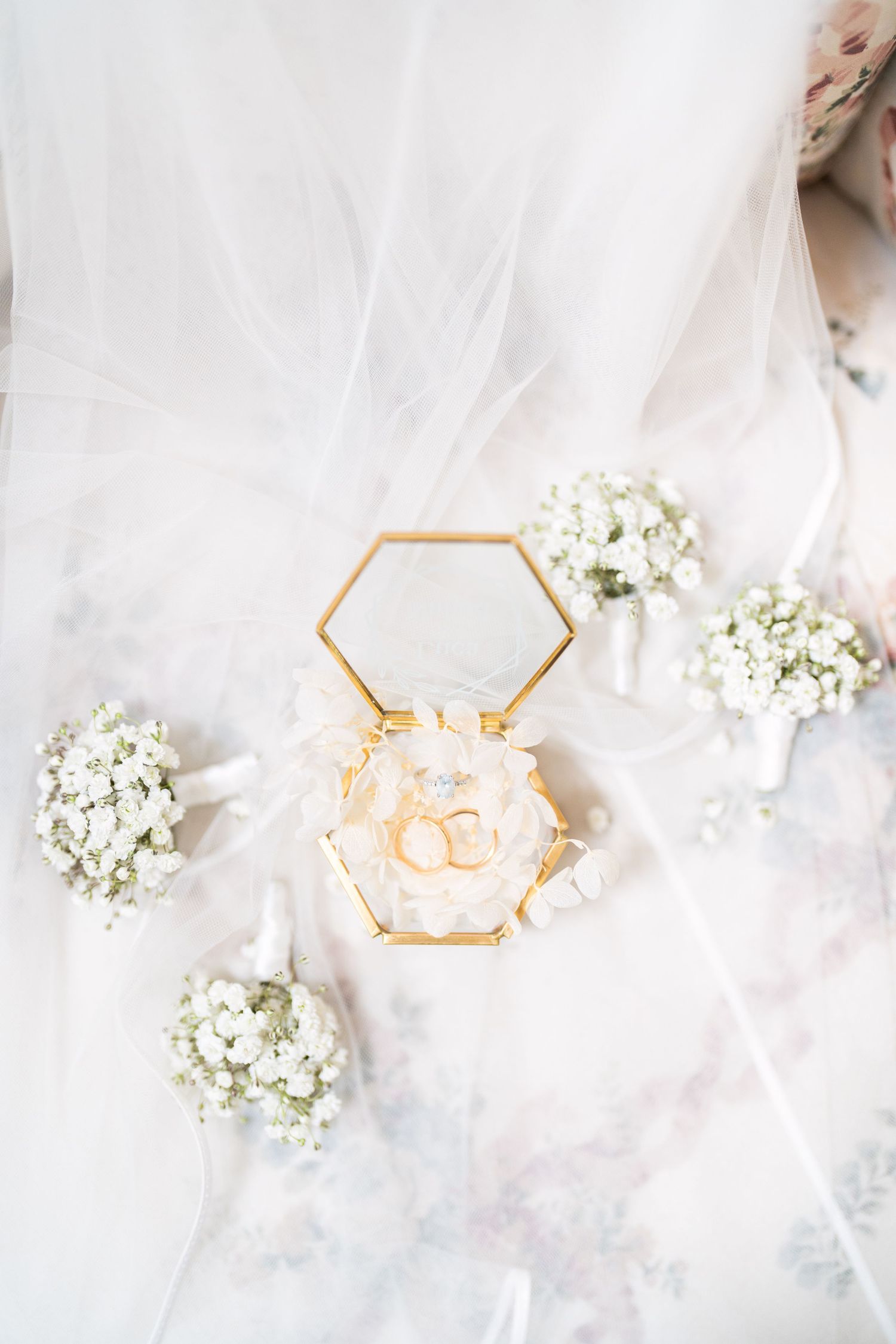 Elegant hexagonal ring box with wedding rings surrounded by delicate baby's breath flowers on white tulle fabric.