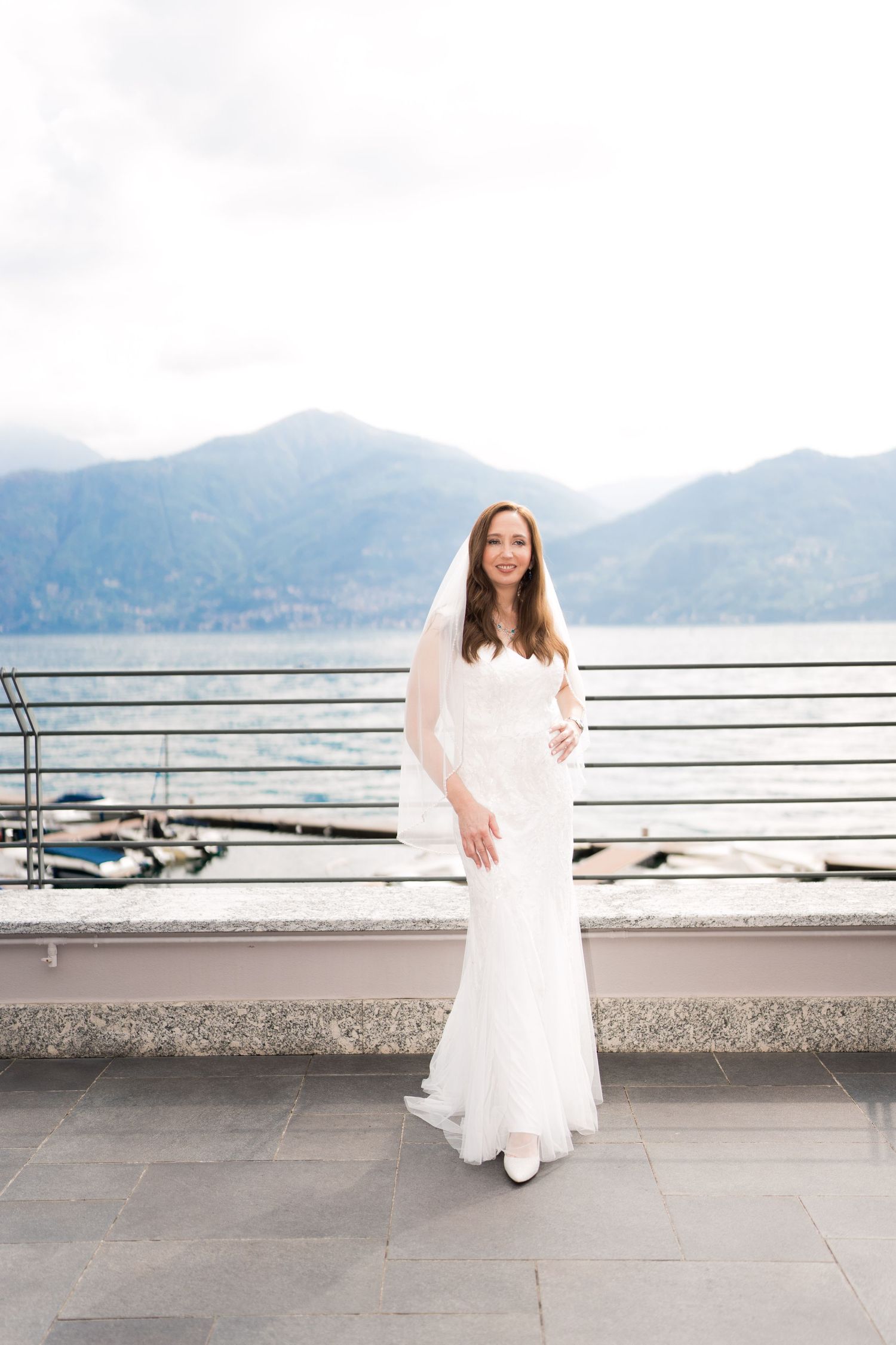 Bride in white gown and veil poses on lakeside terrace with mountain backdrop on her wedding day.