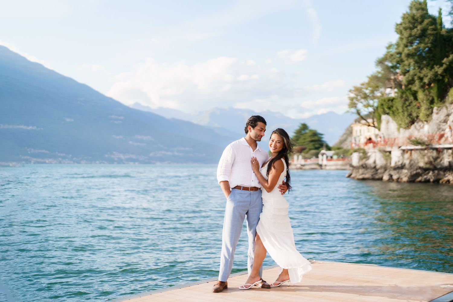 Romantic couple embracing on a lakeside dock with stunning mountain and blue water views in Italy.
