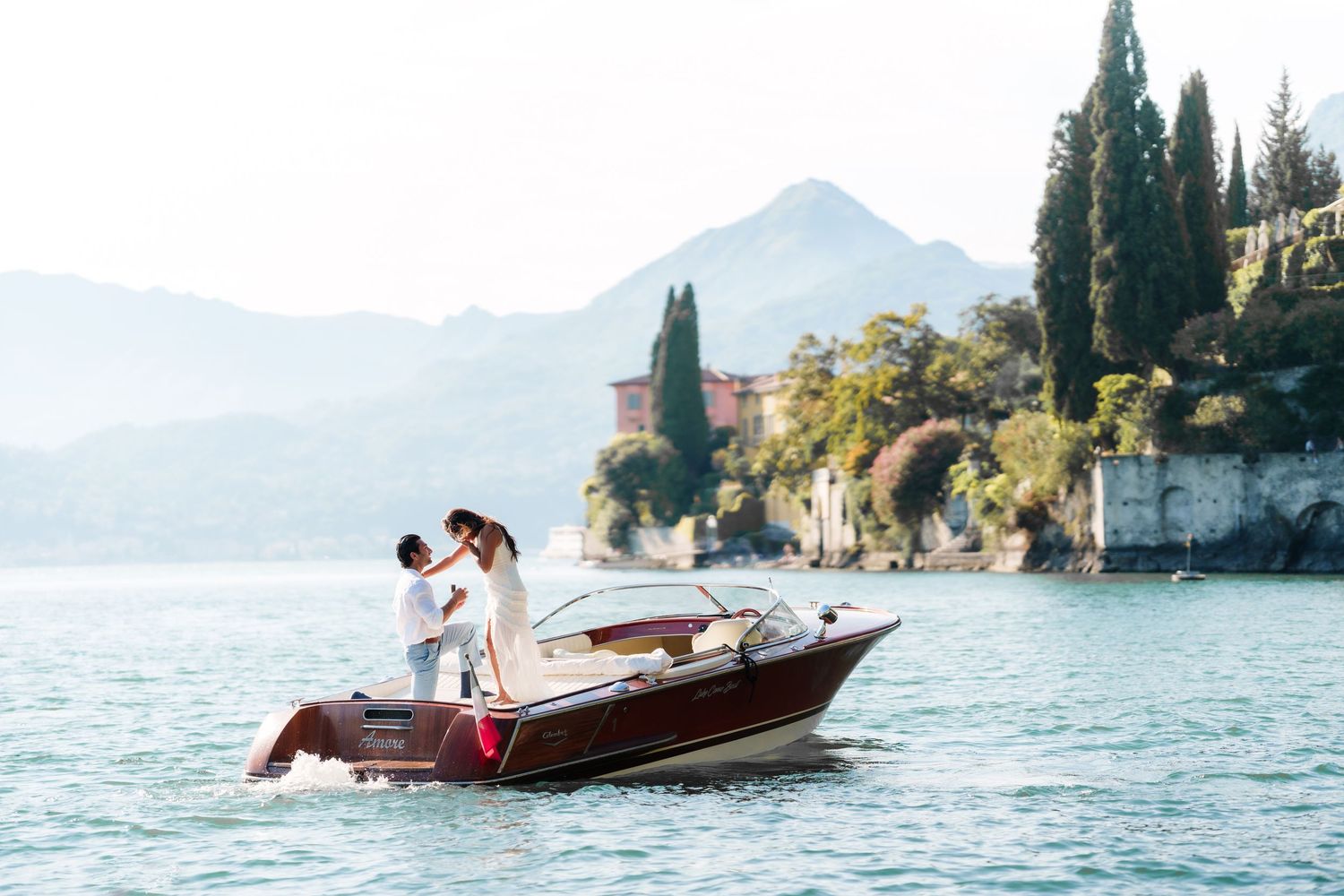 Couple embracing on a classic wooden boat on a serene lake with lush greenery and a historic villa backdrop.