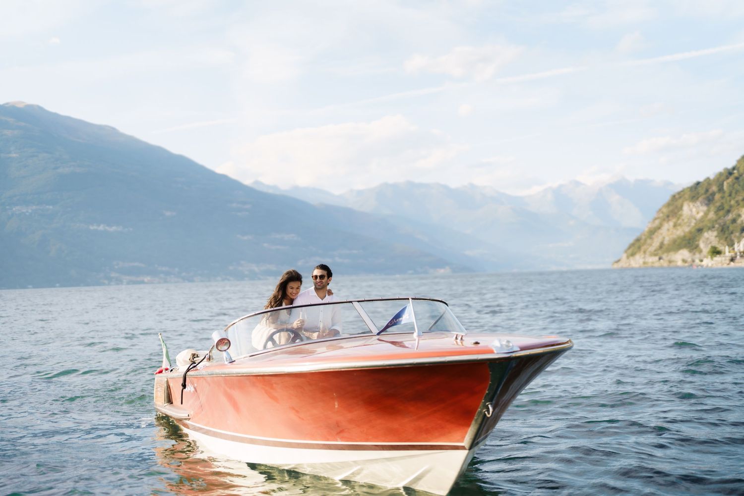 Couple enjoying a romantic boat ride on a serene alpine lake with mountain views on a classic red speedboat.