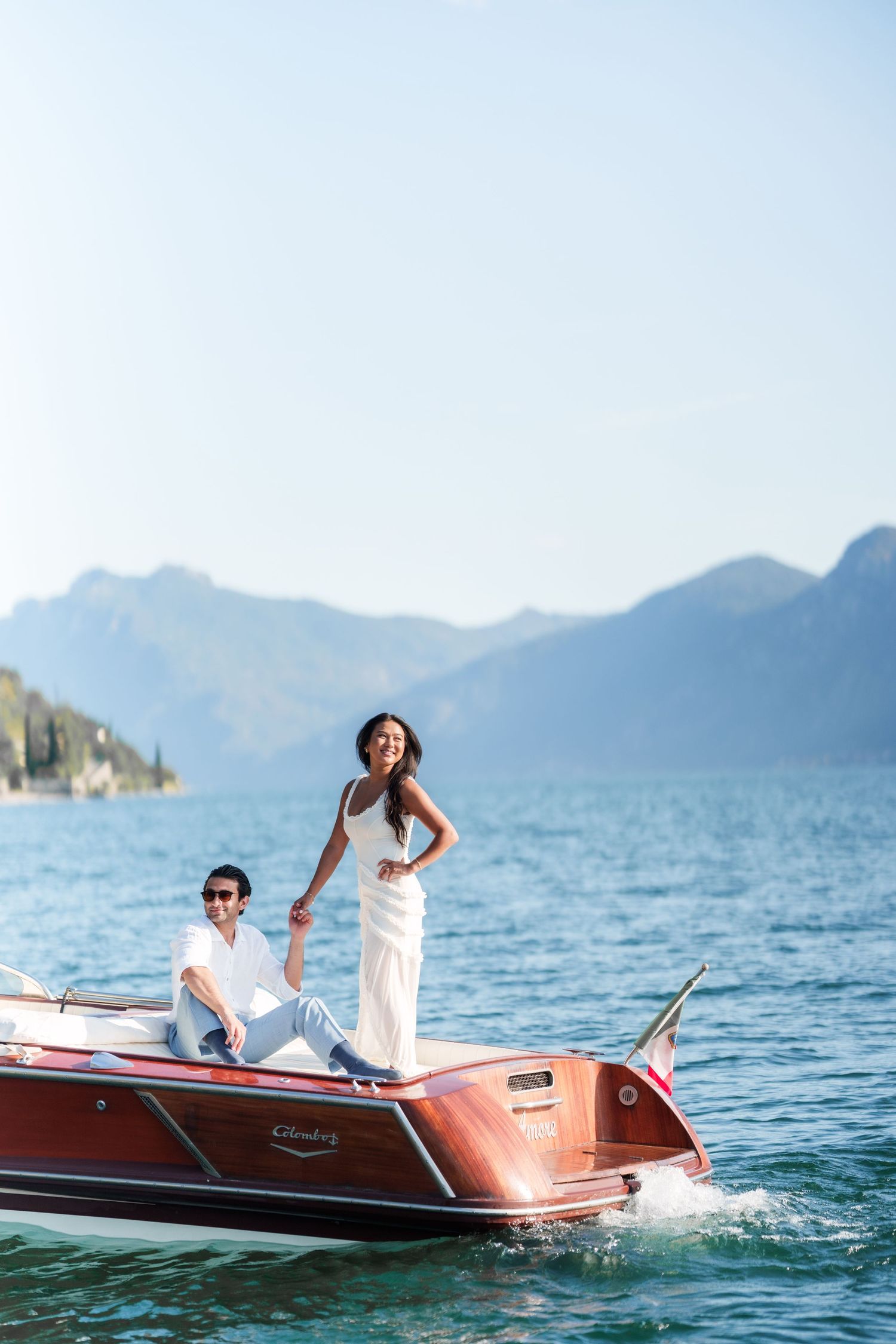 Couple in white outfits on a classic wooden boat on a blue lake with mountain backdrop.