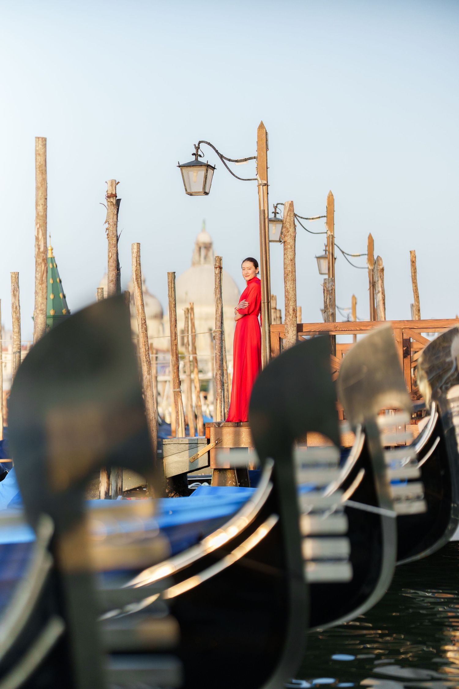 Woman in red dress standing on Venice gondola dock with mooring poles and church in background.