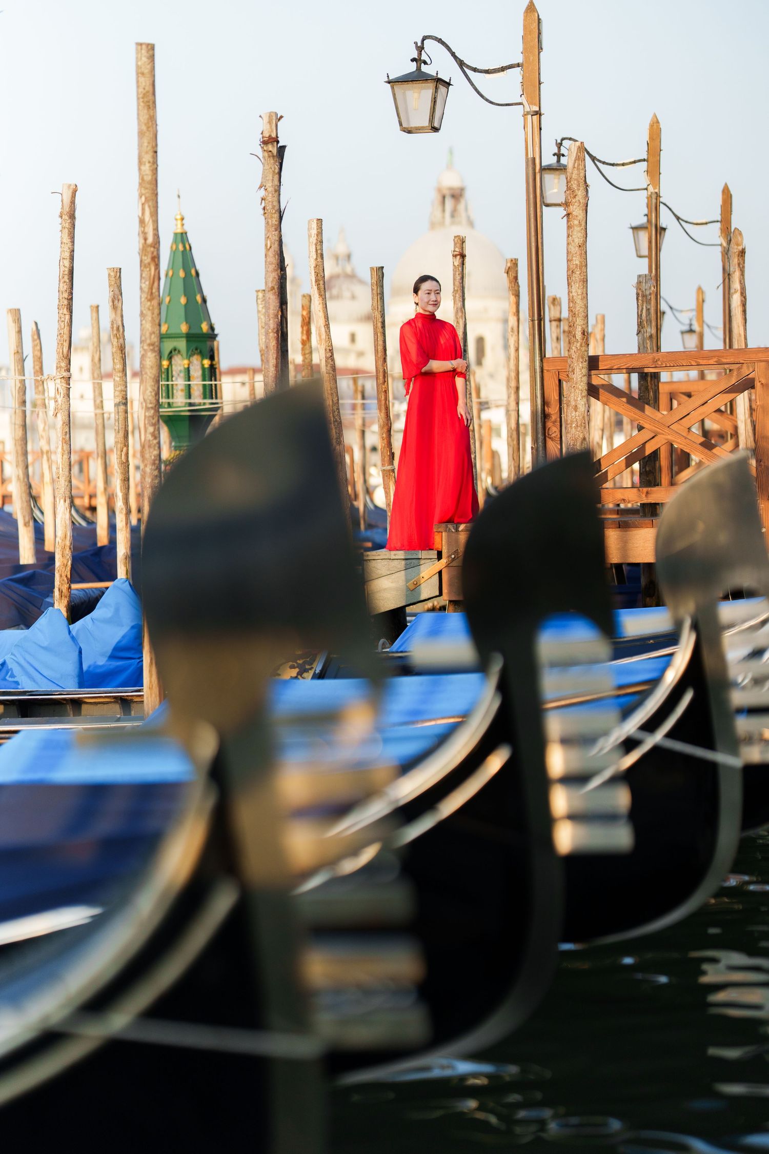 Woman in red dress standing among gondolas at Venice waterfront with San Giorgio Maggiore church in background.