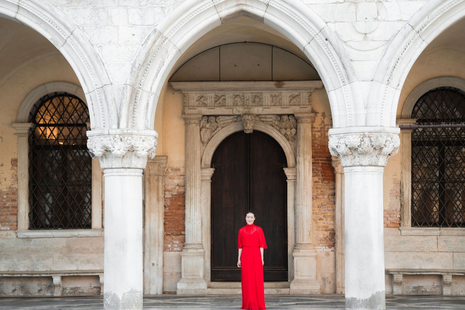 Woman in red dress standing before ornate arched doorway of historic Venetian building with marble columns.