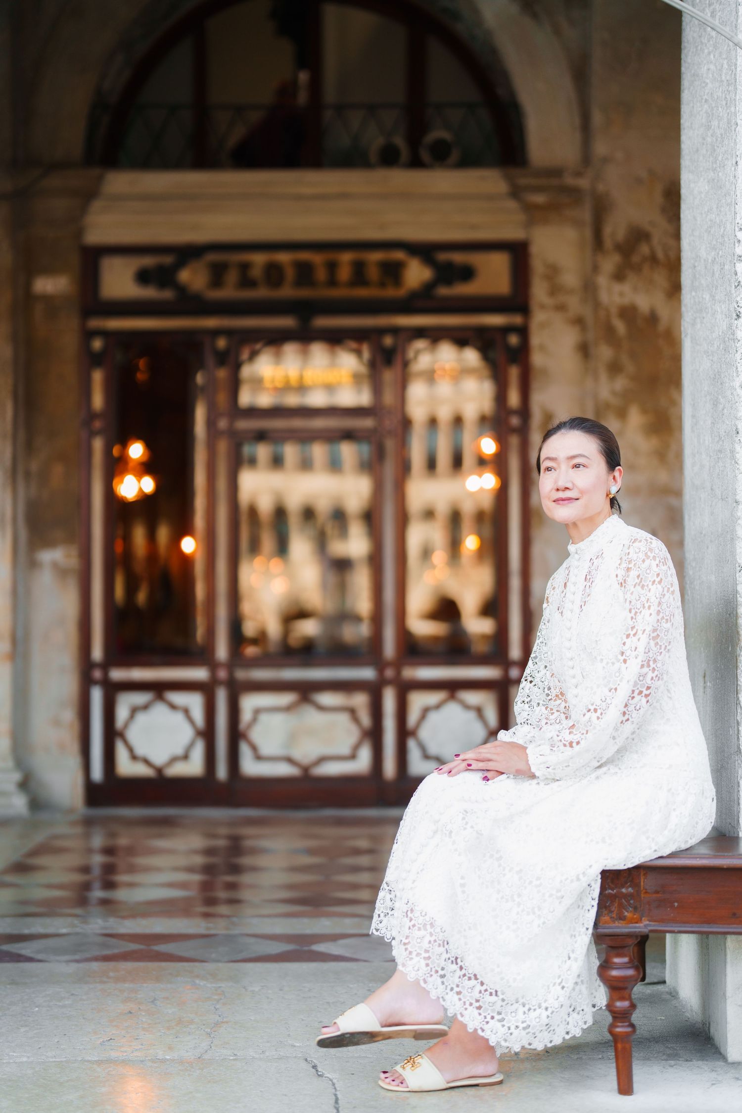 Woman in white lace dress seated outside Caffè Florian in Venice, Italy, smiling gracefully at the camera.