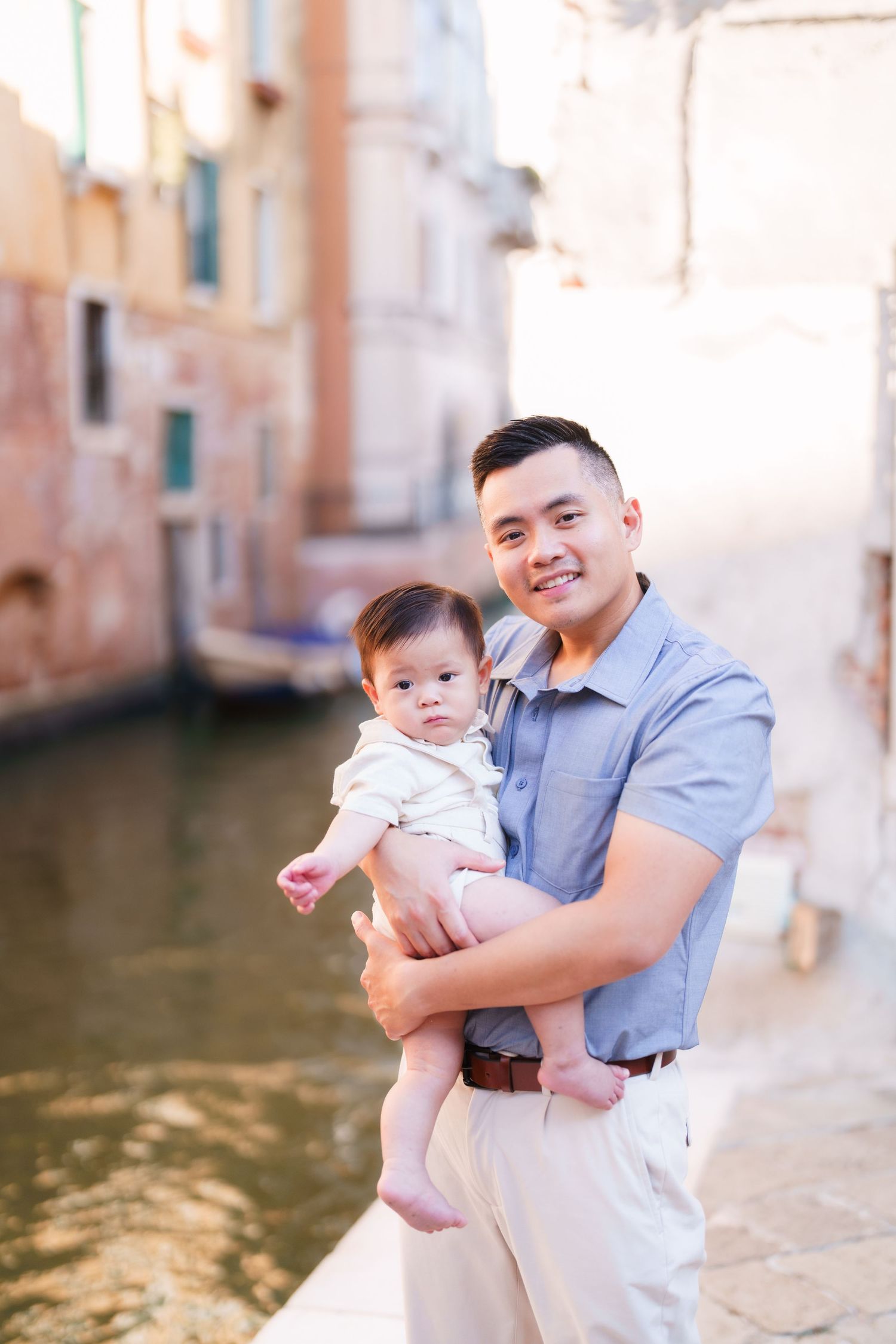 Young Asian father smiling while holding baby in Venice, Italy, with canal and historic buildings in background.