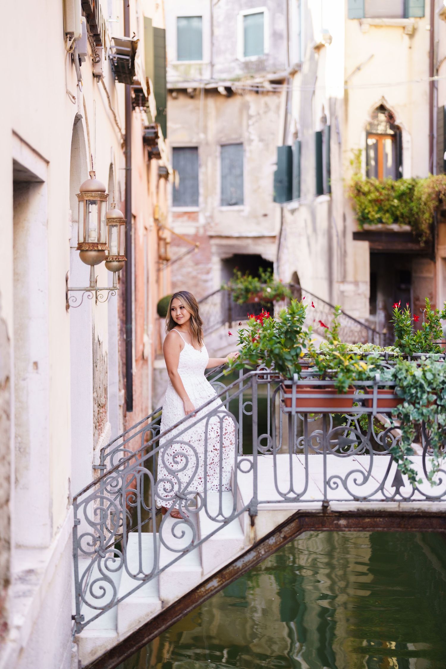 Woman in white dress posing on ornate iron bridge over Venice canal surrounded by lush flowers and historic buildings.