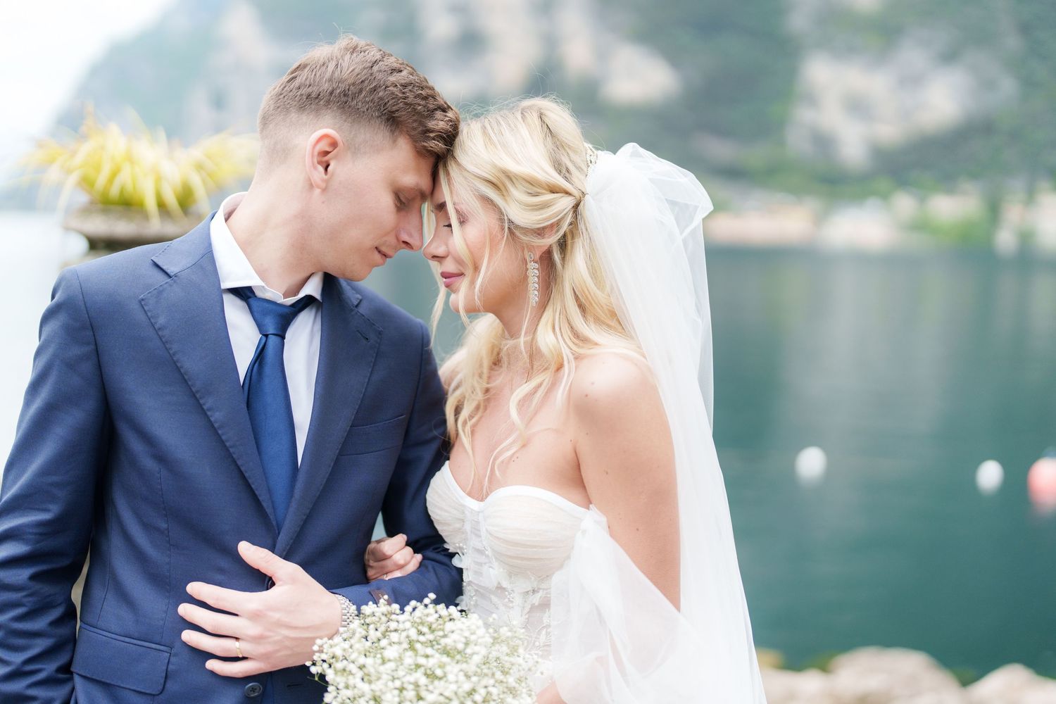 Bride and groom share a romantic forehead touch outdoors, bride holding white flowers, groom in navy suit, lakeside backdrop.