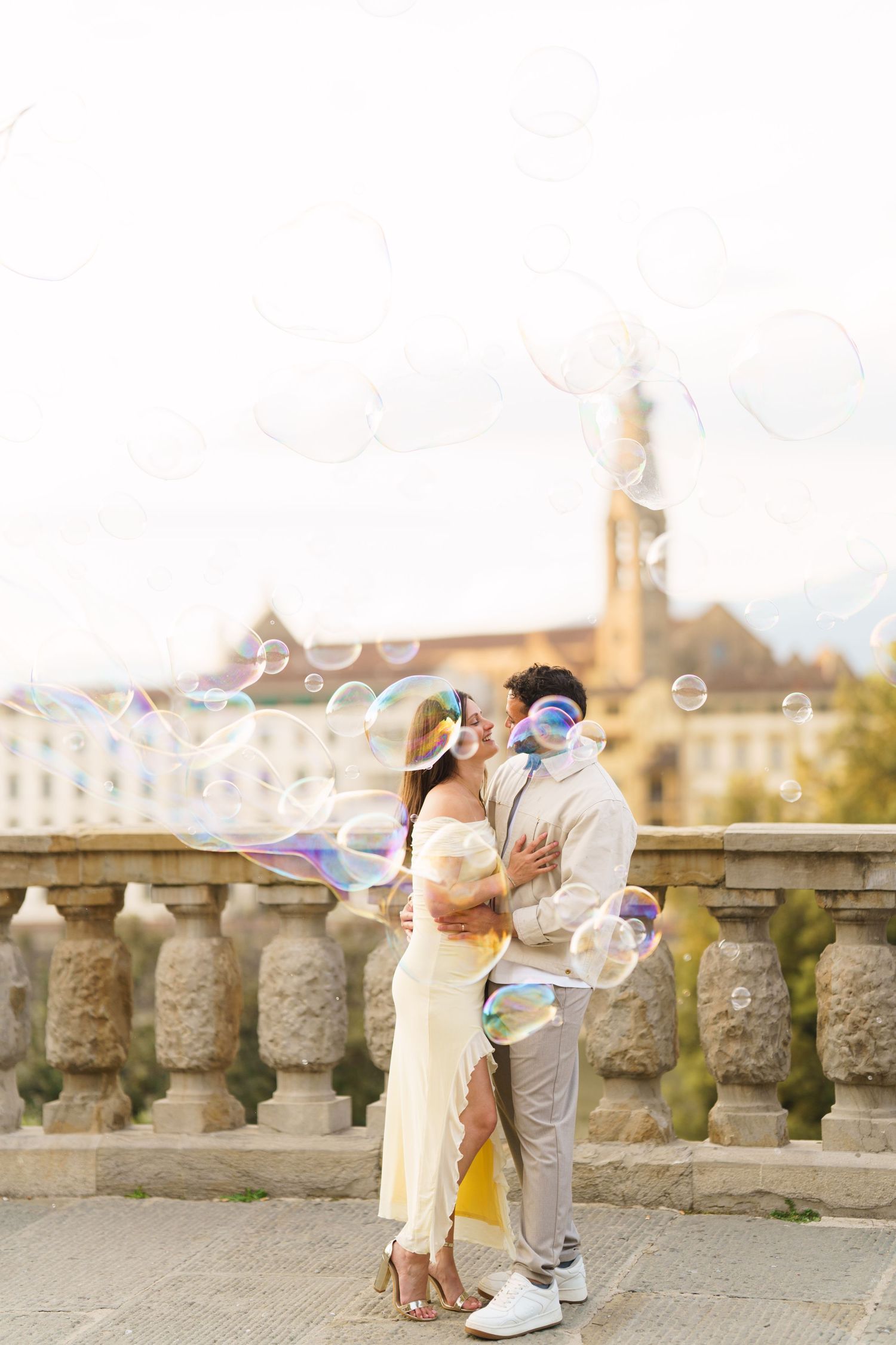 Couple kissing surrounded by soap bubbles on a stone bridge with European architecture in the background.