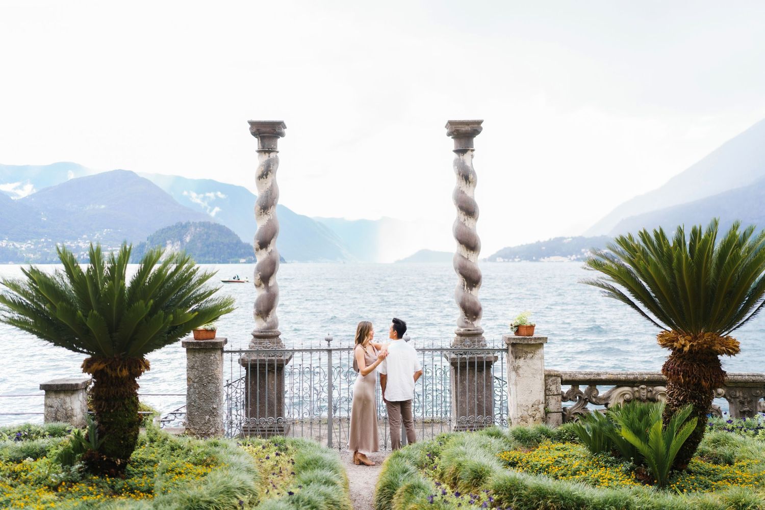 Palm trees and ornate stone columns frame a scenic mountain lake view in an Italian garden setting.