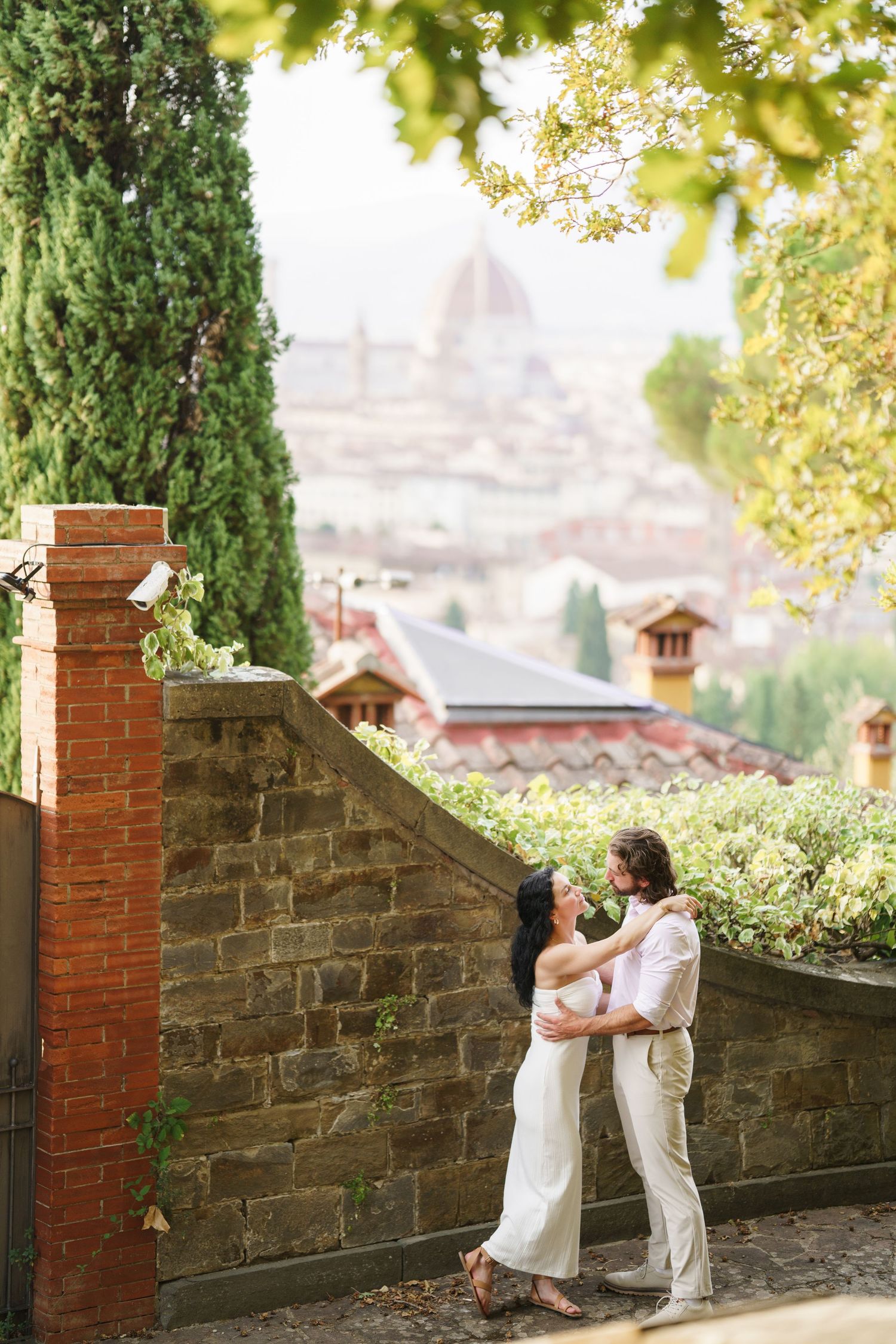 A romantic scene unfolds along a stone wall with cypress trees and golden sunlight filtering through leaves in Florence.