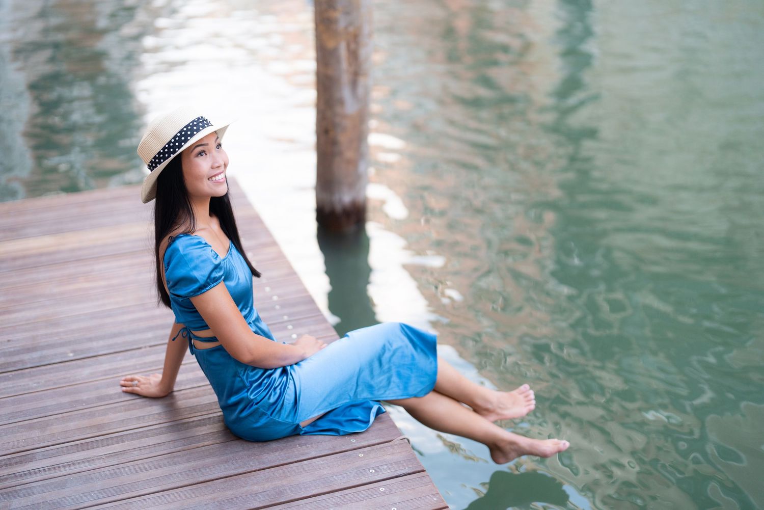 Woman in blue dress and straw hat sits relaxing on wooden dock by tranquil turquoise water.