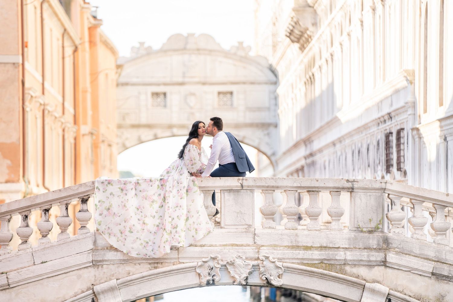 A couple in elegant wedding attire stands on ornate marble steps of a grand historic building in Venice, Italy.