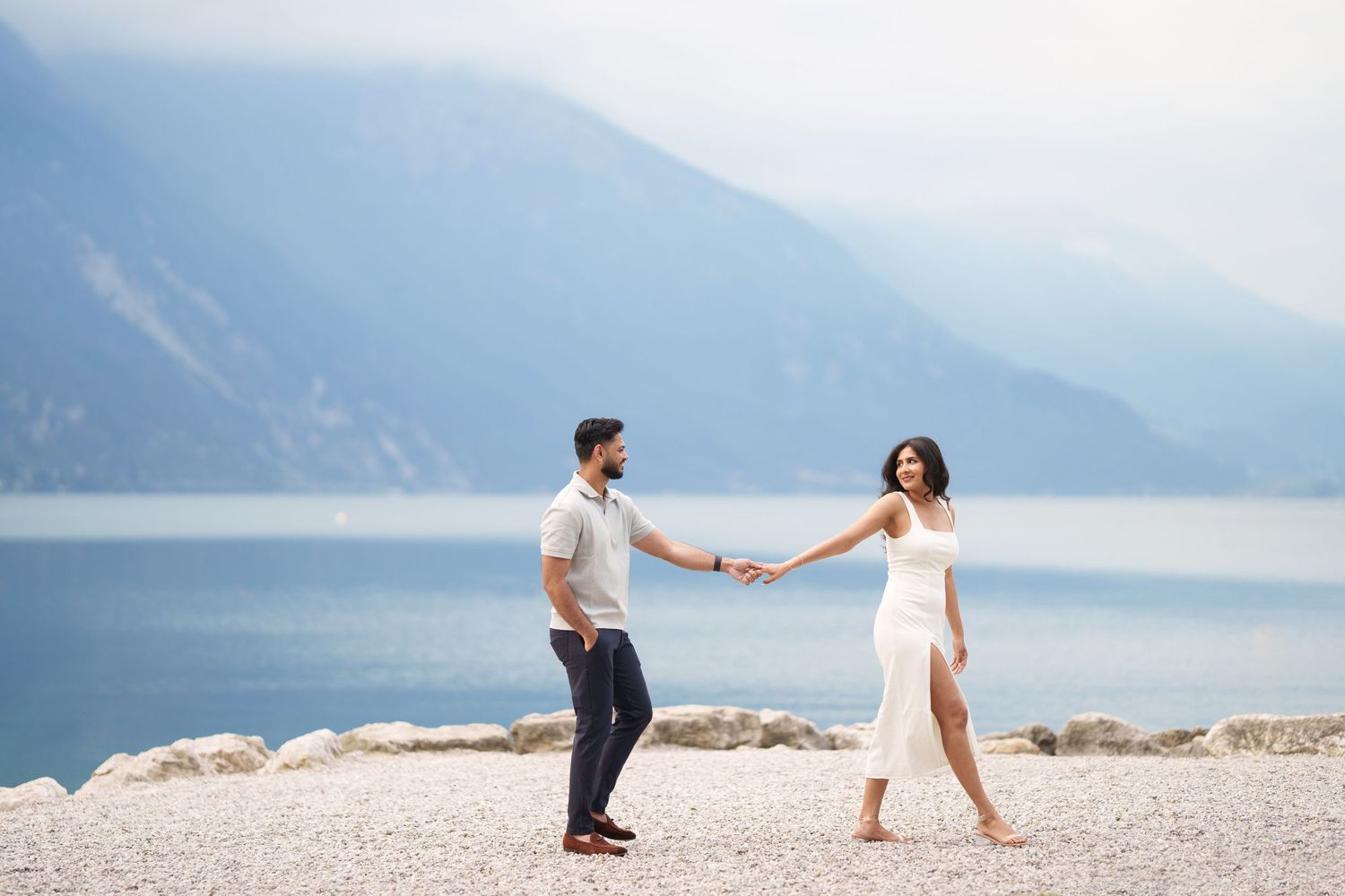 Couple holding hands on rocky beach with misty mountains and lake in background.